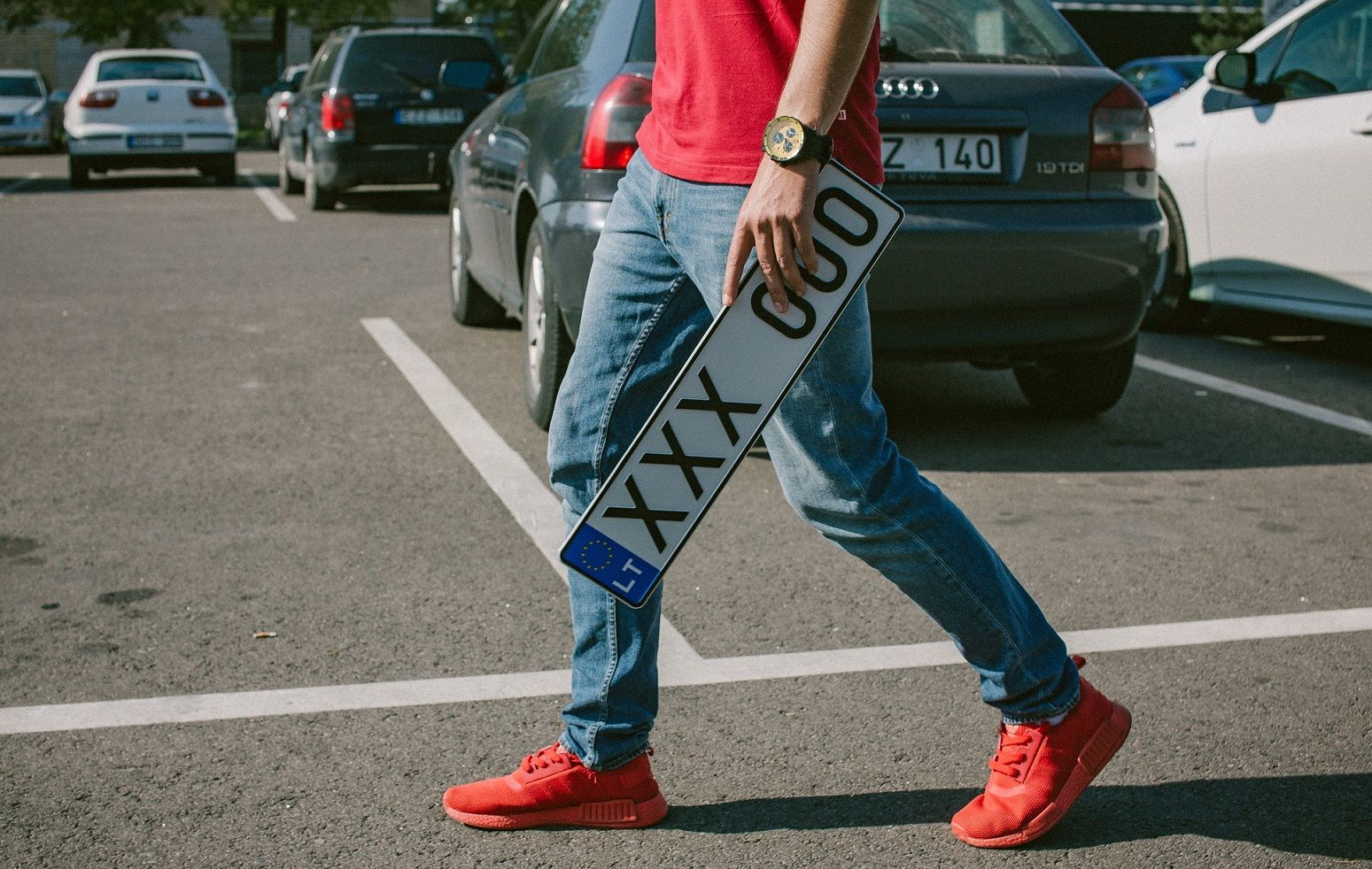 Asmuo su raudonais marškiniais, mėlynais džinsais ir raudonais batais eina per automobilių stovėjimo aikštelę, rankoje laiko europinio stiliaus numerio ženklą „XXX 000“