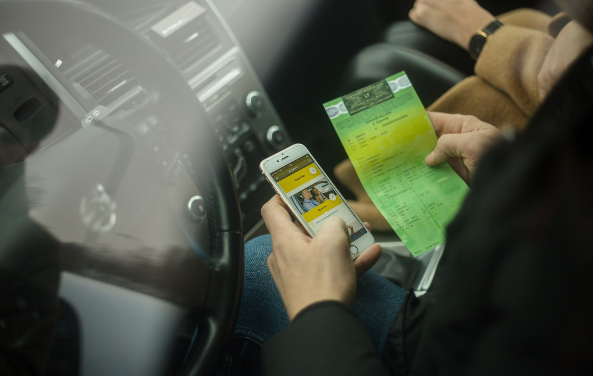 Asmuo automobilyje laiko išmanųjį telefoną ir žalią dokumentą, fone matomas vairas ir prietaisų skydelis