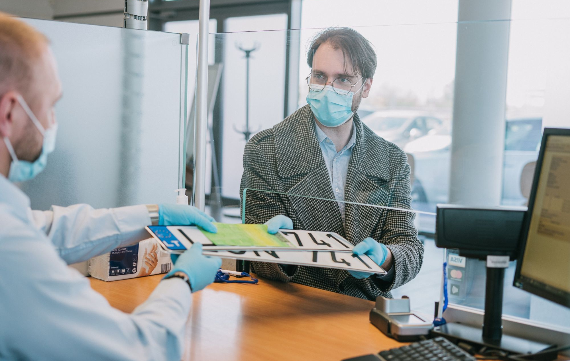 Du žmonės bendrauja prie aptarnavimo langelio, per stiklinę pertvarą perduodami automobilio numerio ženklai ir dokumentai