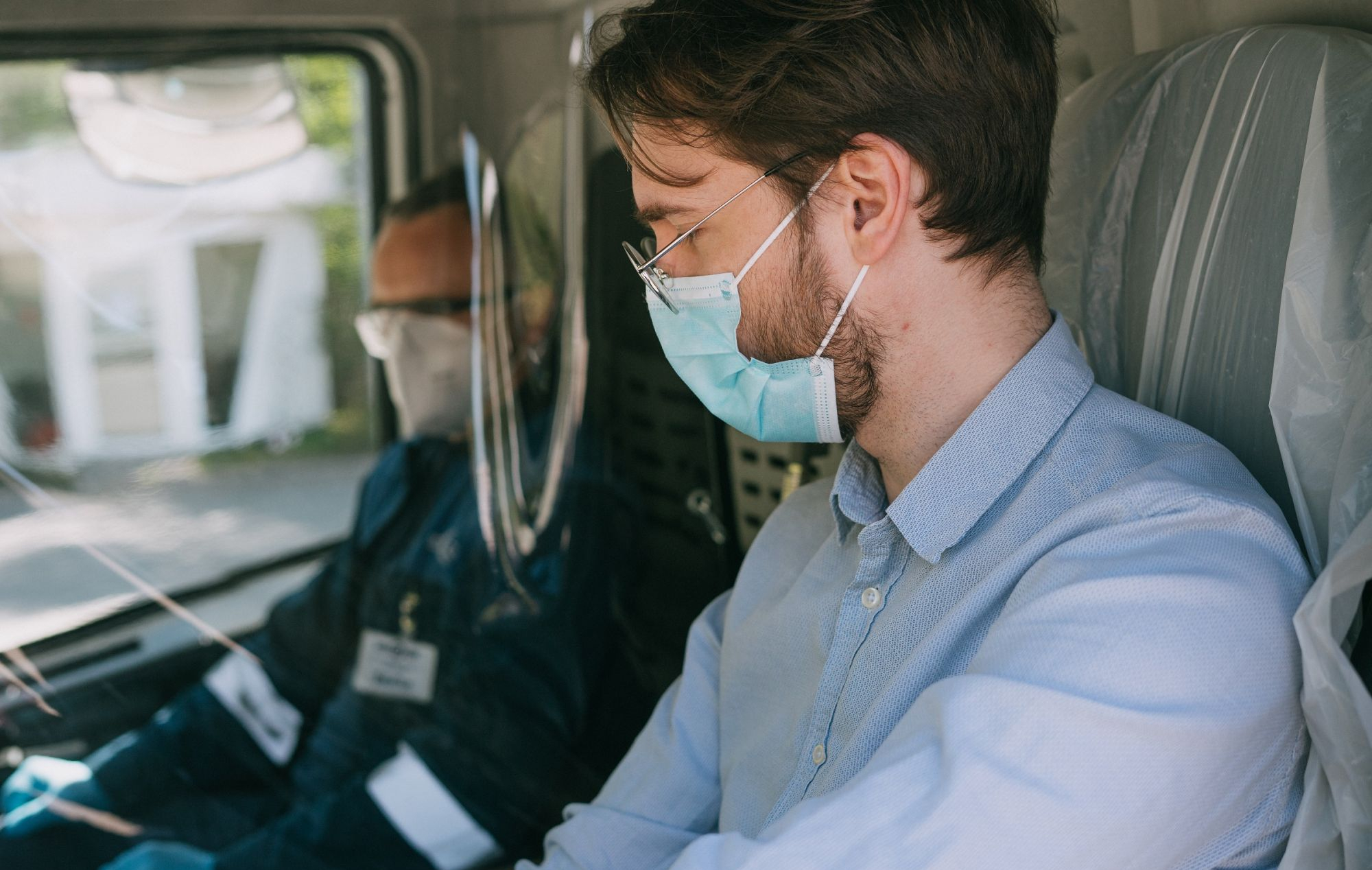 Du žmonės sėdi transporto priemonėje, vienas dėvi apsauginę veido kaukę, pirštines ir turi identifikacinį pažymėjimą