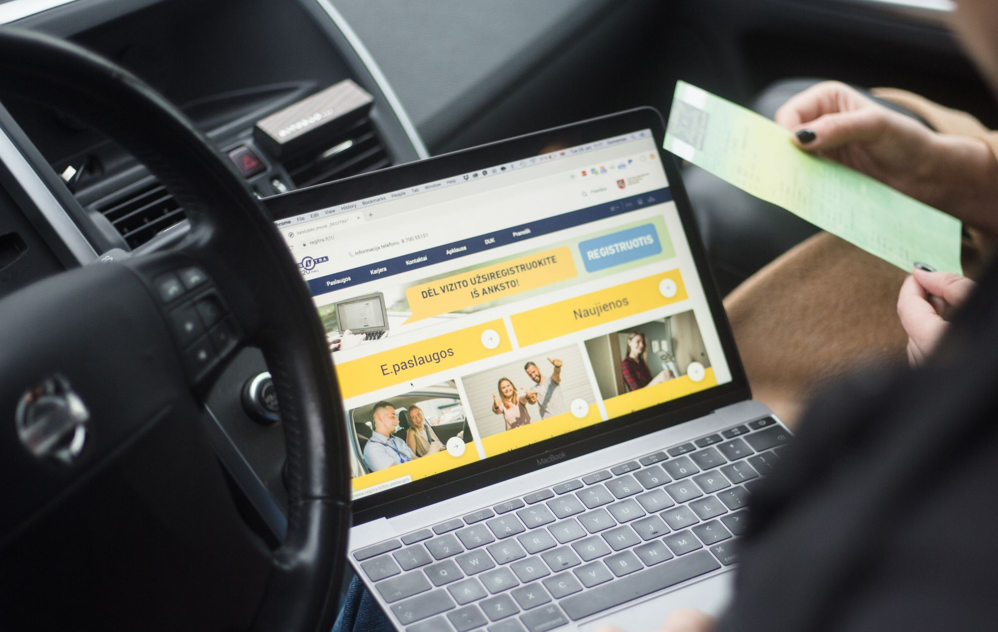 Asmuo automobilyje laiko žalią lapą ir naudojasi nešiojamuoju kompiuteriu, kurio ekrane matoma svetainė su skiltimis „E.paslaugos“ ir „Naujienos“