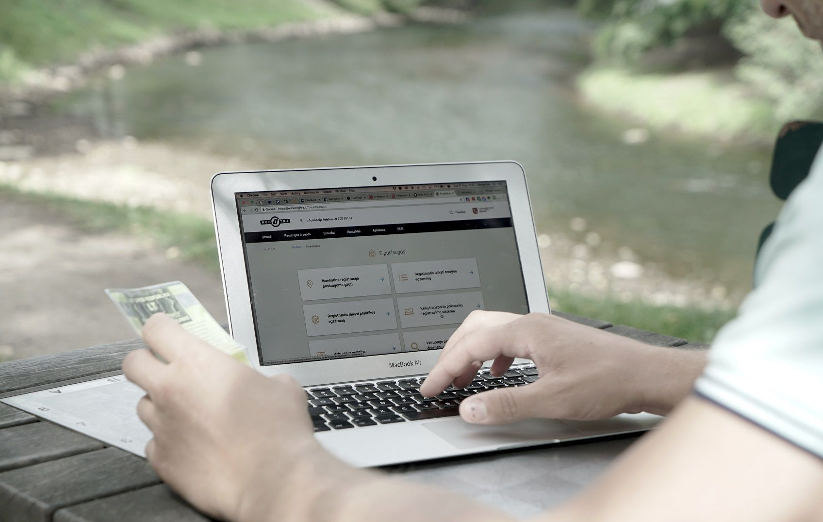 Asmuo lauke prie upės naudoja „MacBook Air“ kompiuterį, vienoje rankoje laikydamas banknotą