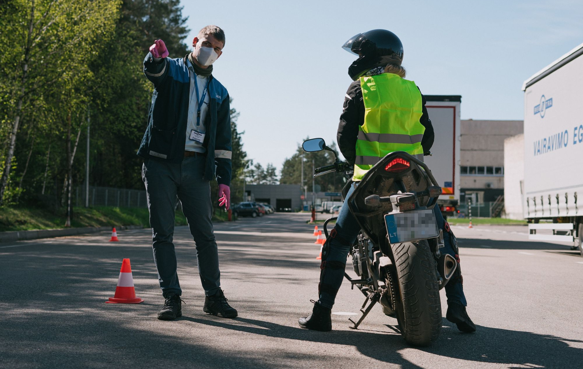 Motociklininkas su šalmu ir ryškiaspalve liemene mokomas instruktoriaus šalia kelio su eismo kūgiais