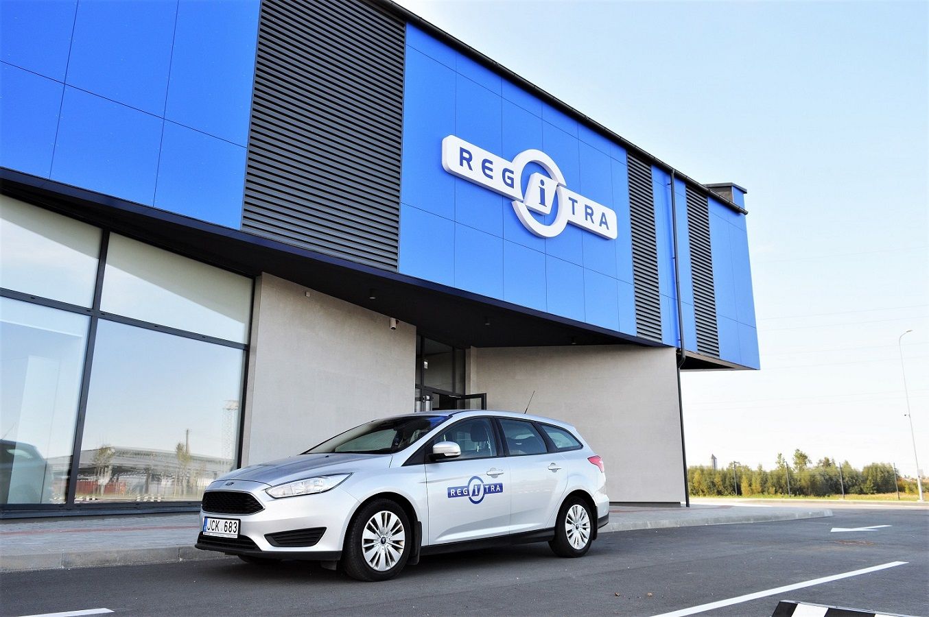 Pastatas mėlynu fasadu su užrašu „REGITRA“, prie jo stovi sidabrinis automobilis su „REGITRA“ logotipu