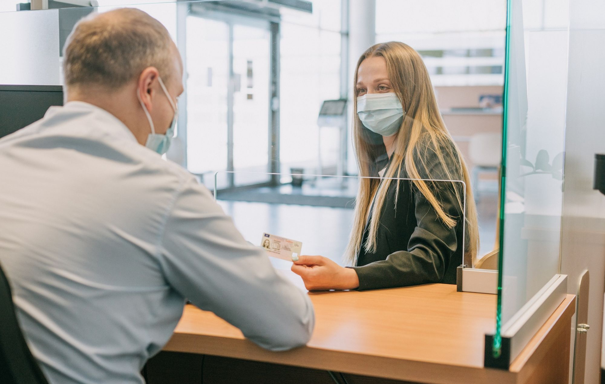 Du žmonės sėdi priešais vienas kitą prie stalo biure, vienas perduoda asmens tapatybės kortelę per stiklinę pertvarą