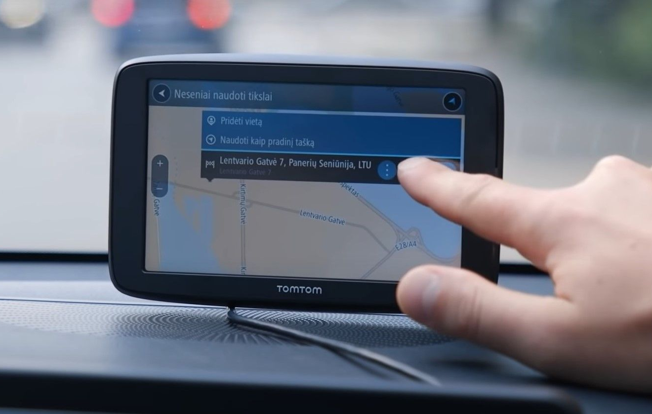 GPS navigacijos įrenginys automobilyje rodo žemėlapį su pažymėta vieta „Lentvario gatvė 7“, ranka rodo į ekraną