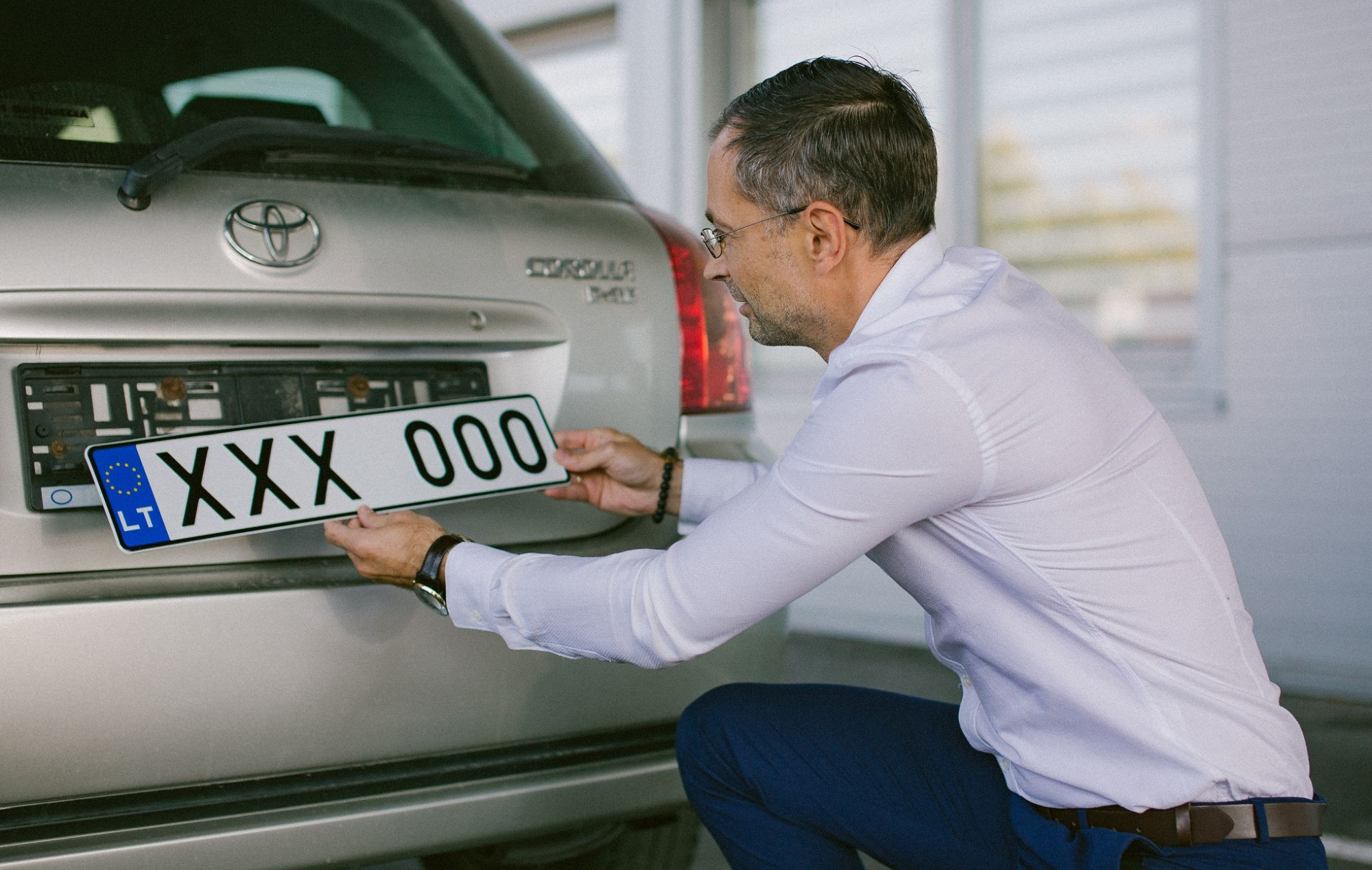 Asmuo tvirtina lietuvišką numerio ženklą „XXX 000“ prie sidabrinio „Toyota“ automobilio galo.