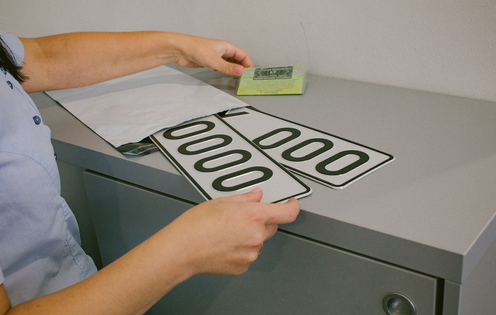 Asmuo prie pilkos spintelės laiko du stačiakampius objektus su užrašu „0000“, ant spintelės padėta žalia kortelė