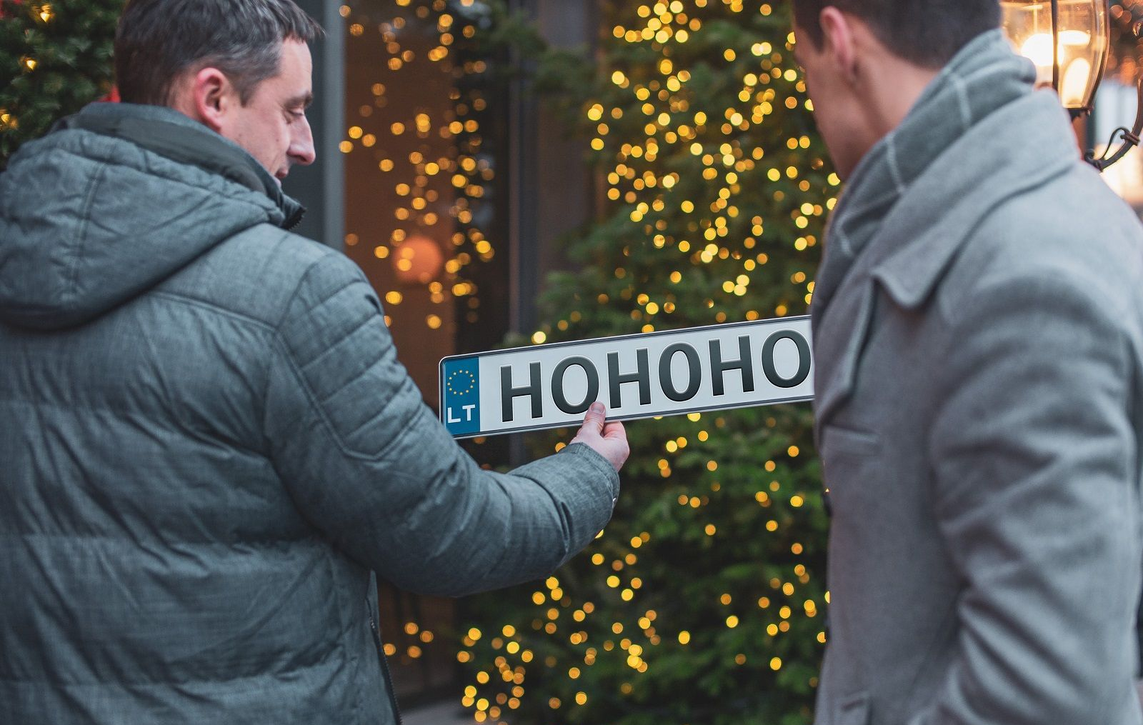 Du žmonės lauke, vienas laiko dekoratyvinį numerio ženklą su užrašu „HOHOHO“, fone – kalėdinė eglutė su girliandomis