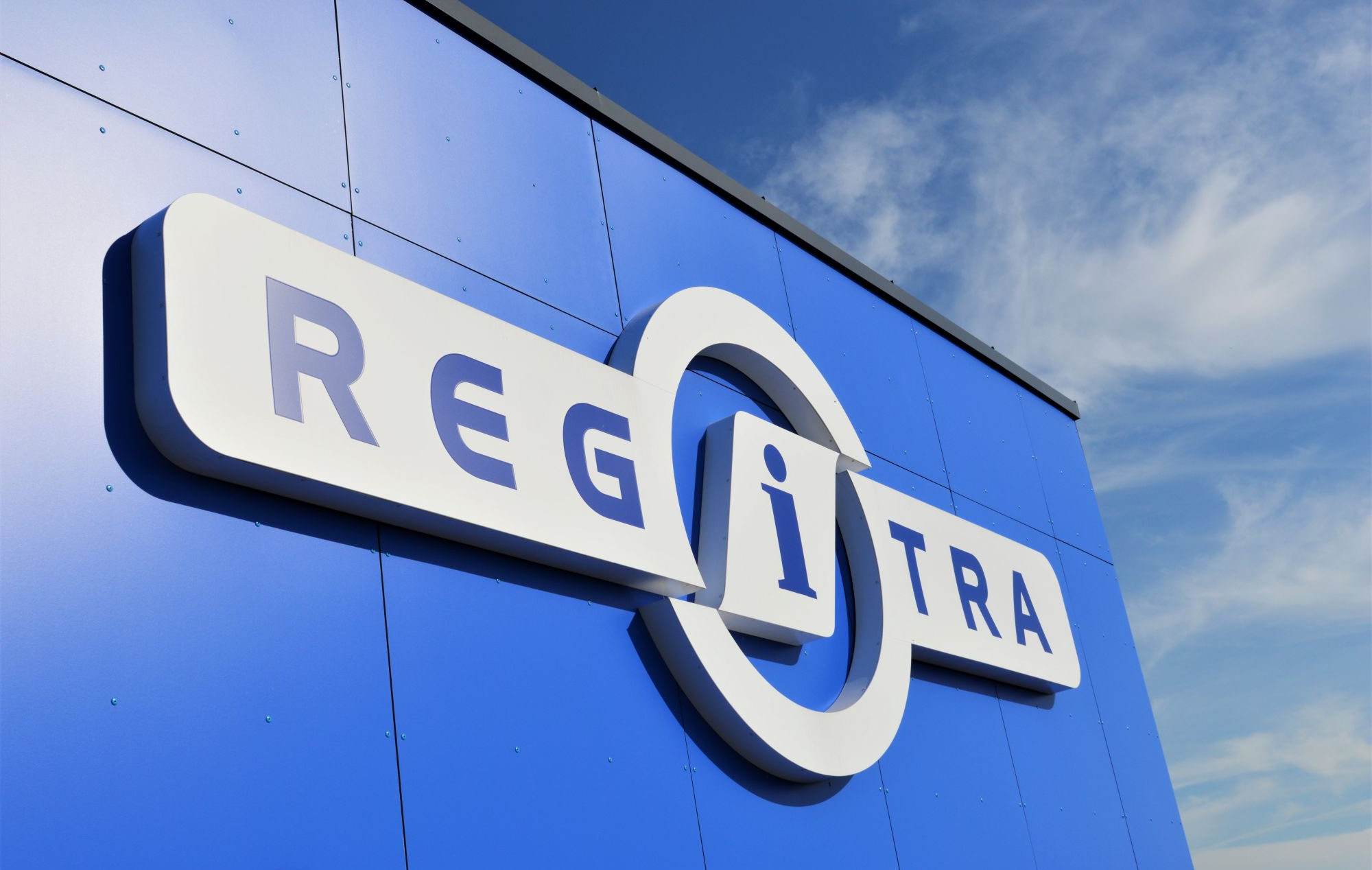 Mėlynas pastatas su baltu užrašu „REGITRA“ ir logotipu, fone matomas dangus su debesimis