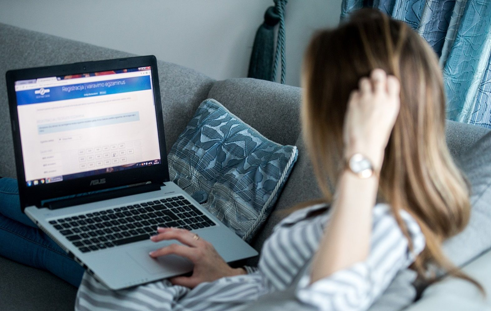 Asmuo sėdi ant sofos, naudojasi ASUS nešiojamuoju kompiuteriu, kurio ekrane matoma svetainė „Registracija ir vairavimo egzaminai