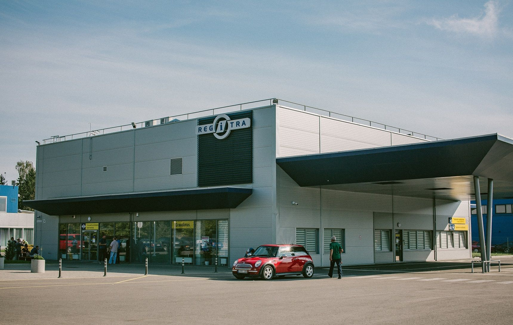 Modernus pastatas su užrašu „REGITRA“, prie įėjimo stovi žmonės, o priekyje – raudonas automobilis.