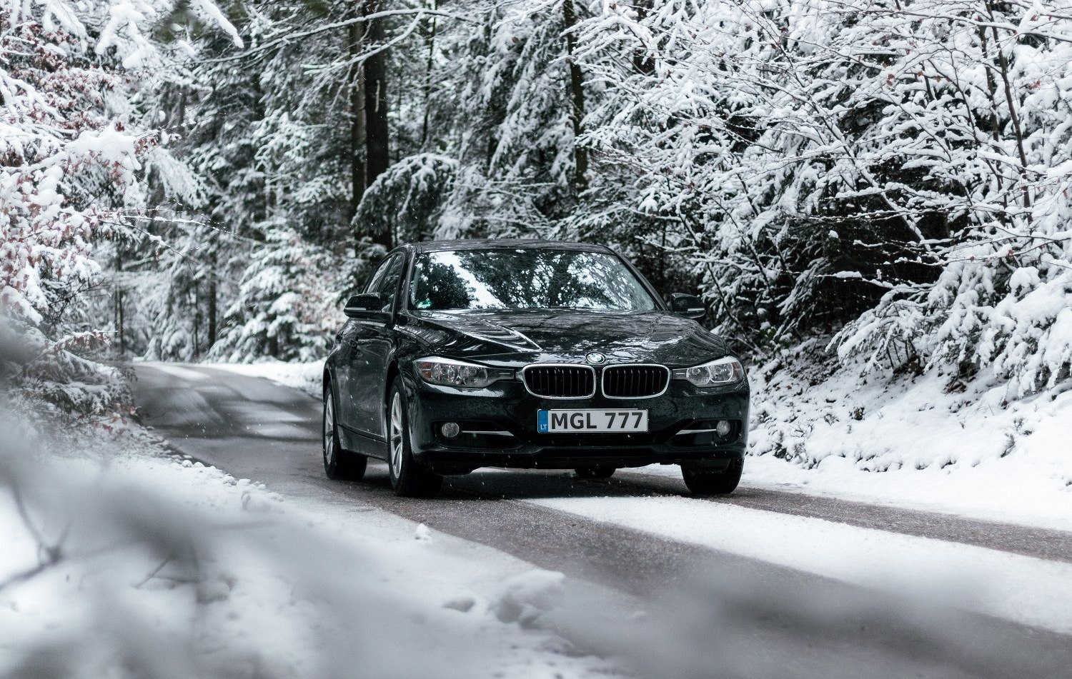 Juodas BMW automobilis su numeriu „MGL 777“ važiuoja snieguotu miško keliu žiemiškoje aplinkoje.
