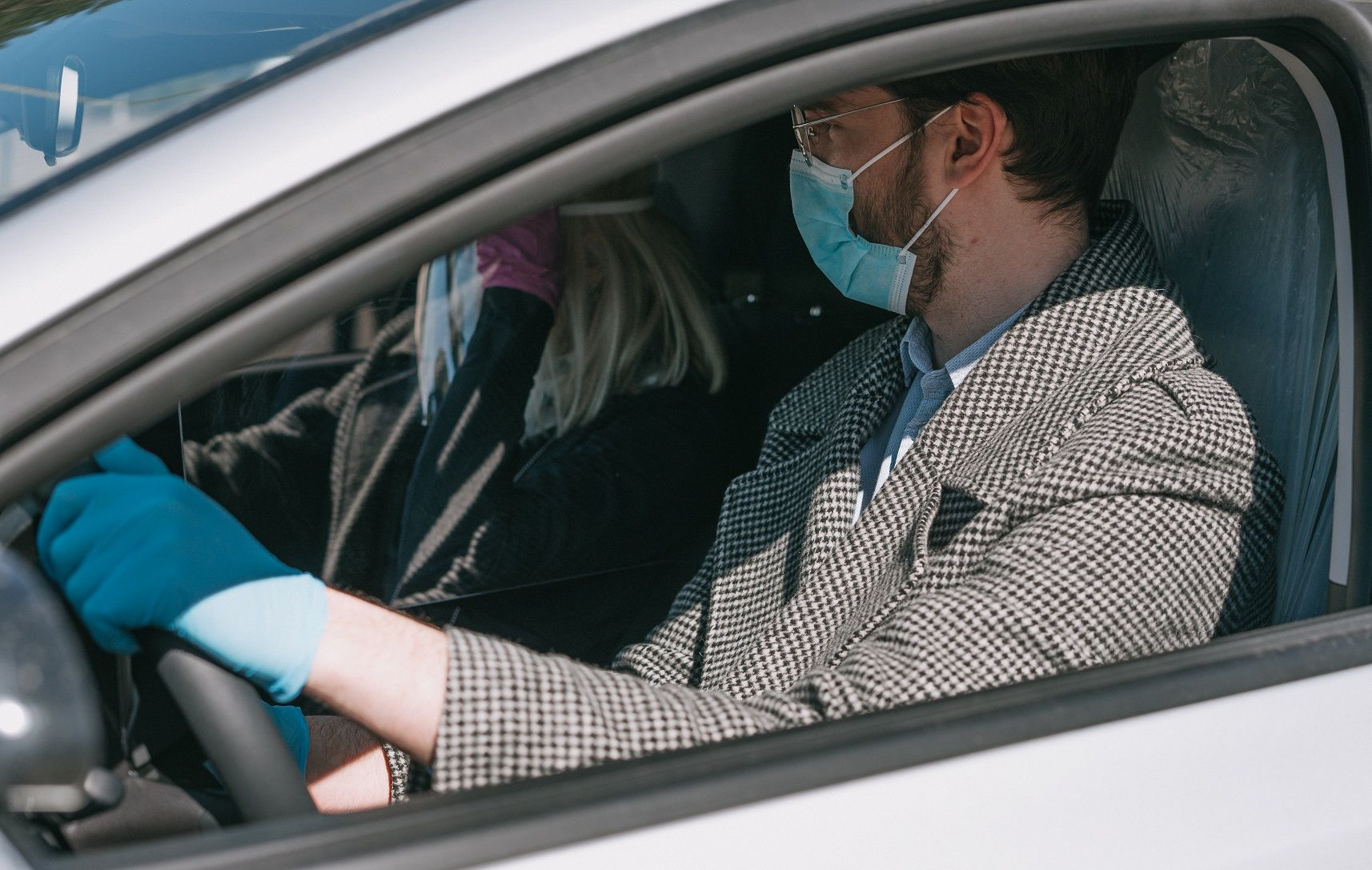 Du asmenys automobilyje – vairuotojas su languotu paltu, mėlynomis pirštinėmis ir kauke, keleivis taip pat su kauke ir pirštinėmis.