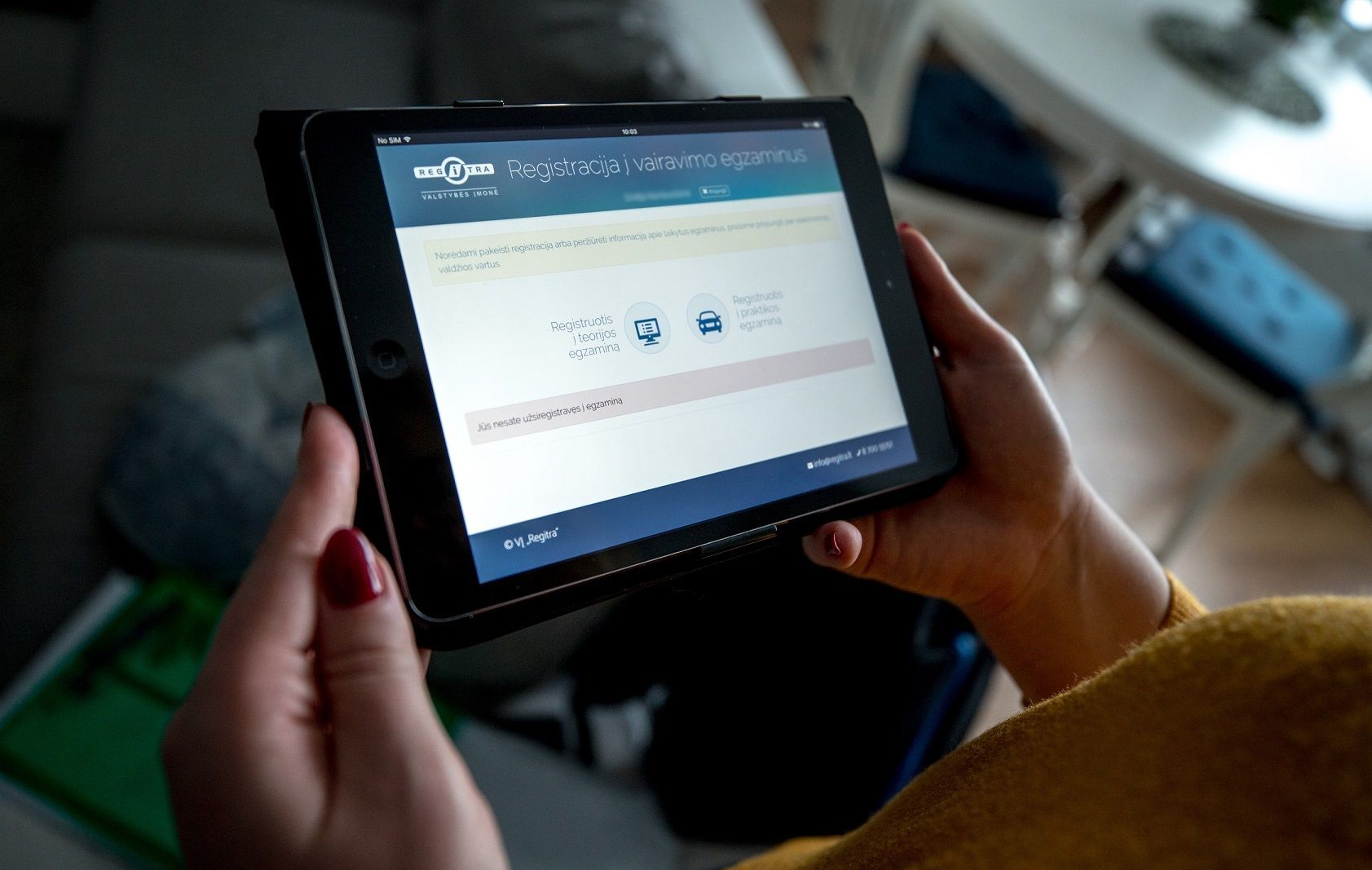 Asmuo laiko planšetę, kurioje atidaryta svetainė „Registracija į vairavimo egzaminus“ su pasirinkimais teorijos ir praktikos egzaminams.