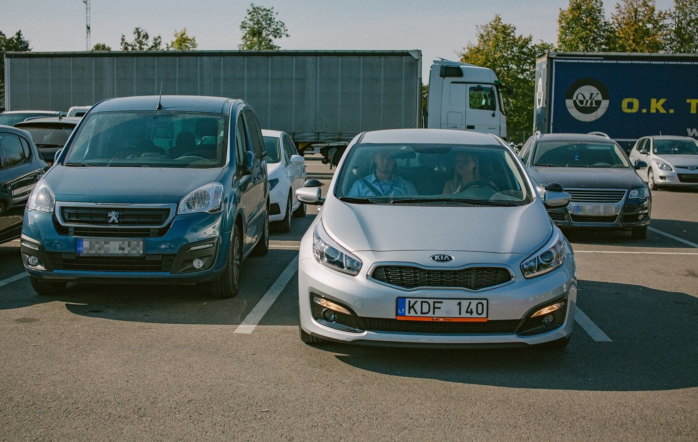 Mėlynas „Peugeot“ ir sidabrinis „Kia“ automobiliai stovėjimo aikštelėje, „Kia“ numeris – „KDF 140“, fone matomas sunkvežimis ir transporto priemonė su užrašu „O.K.“.