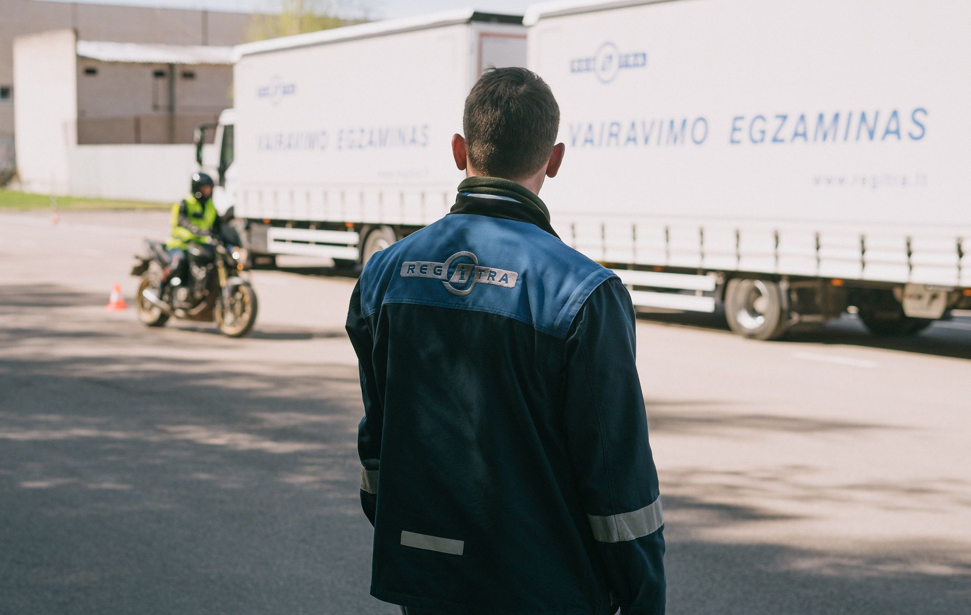 Asmuo su „REGITRA“ striuke stebi motociklininką prie dviejų sunkvežimių su užrašais „VAIRAVIMO EGZAMINAS“ ir „www.regitra.lt“.