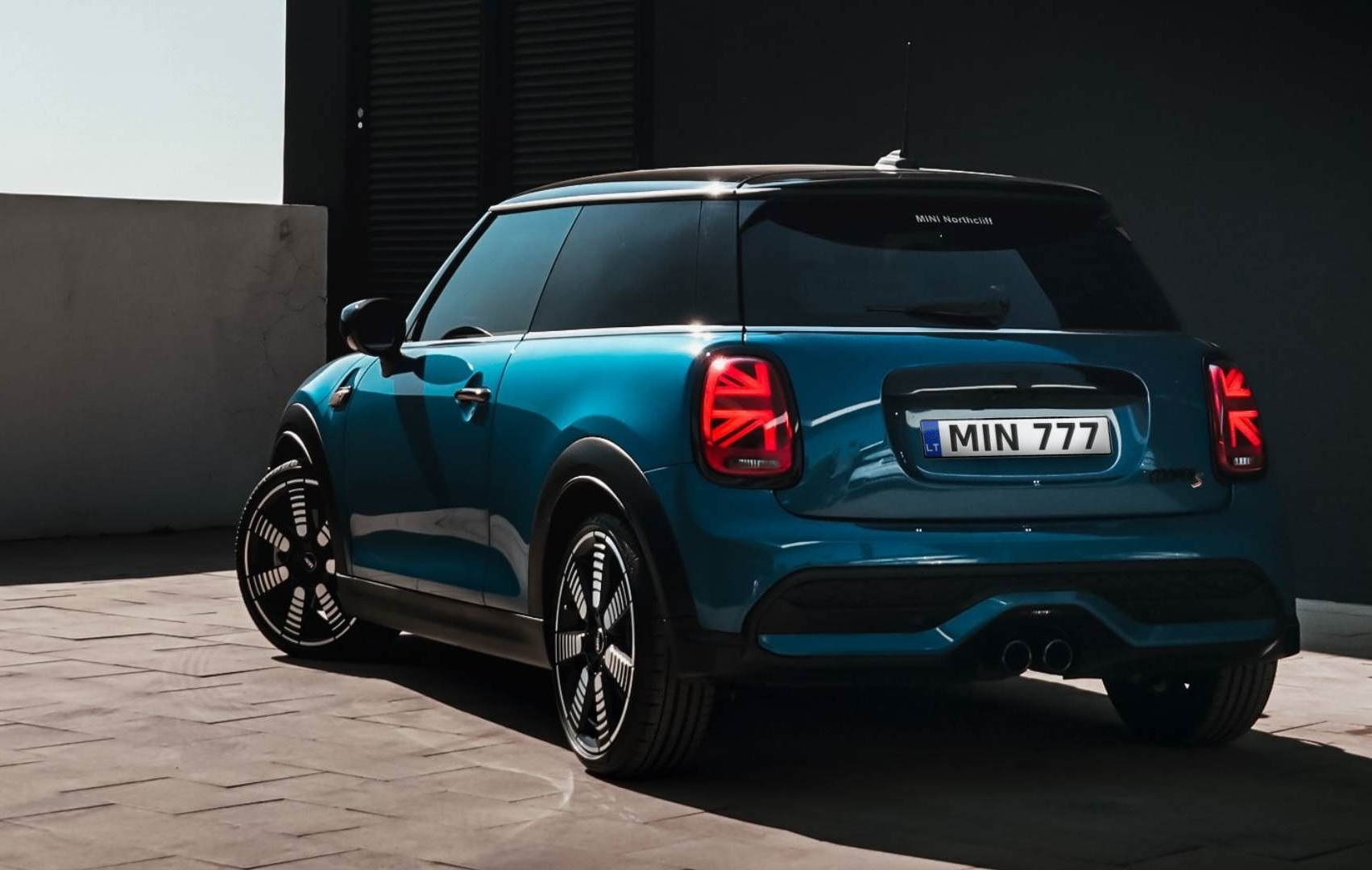 Mėlynas „Mini Cooper“ su juodos spalvos stogu, „Union Jack“ dizaino galiniais žibintais ir numeriu „MIN 777“, stovintis ant trinkelių