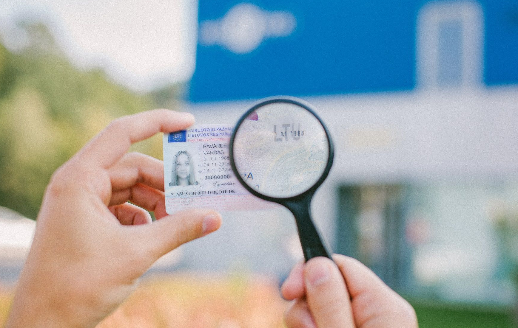 Asmuo laiko Lietuvos asmens tapatybės kortelę ir padidinimo stiklą, fone matoma žaluma ir mėlynas pastatas.
