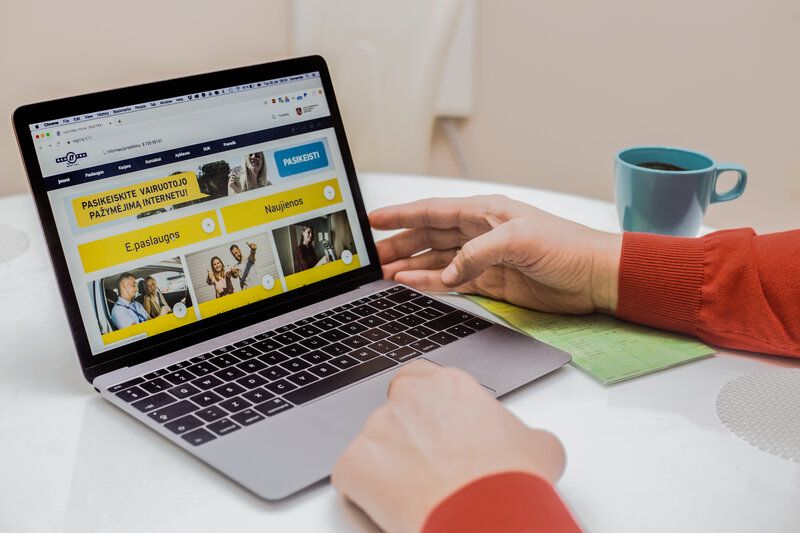 Asmuo naudojasi nešiojamuoju kompiuteriu, ekrane matoma svetainė su tekstu „PASIRINKITE VAIRUOTOJO PAŽYMĖJIMĄ INTERNETUI“, šalia mėlynas puodelis ir dokumentai.