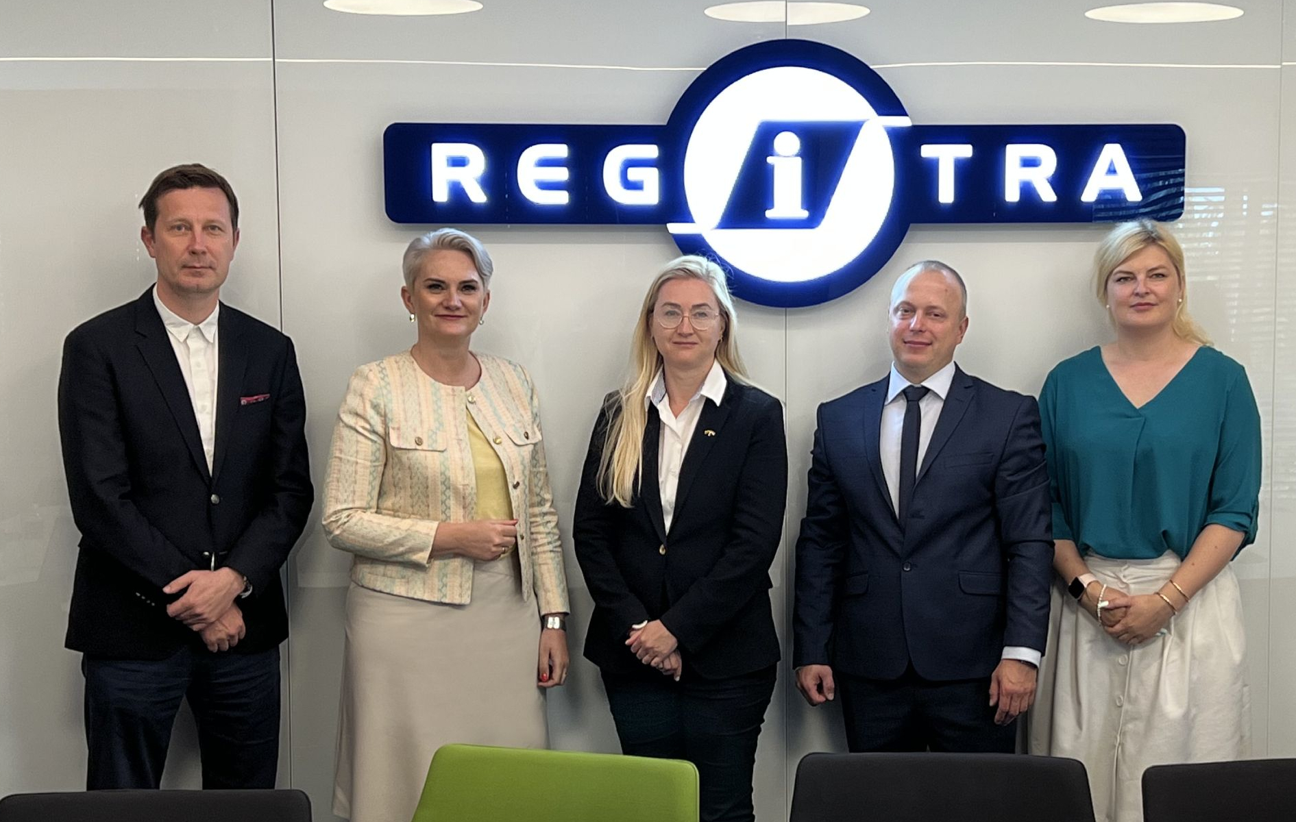 Penki žmonės stovi prieš sieną su „REGITRA“ logotipu, jų veidai užbluruoti.