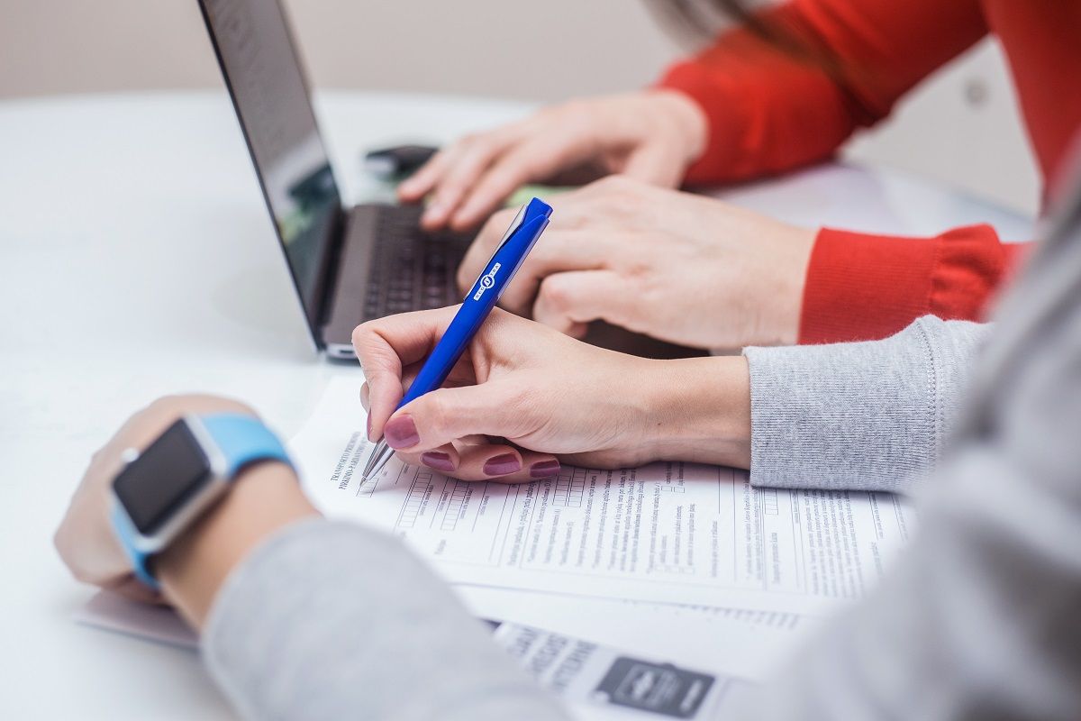 Du žmonės dirba prie stalo – vienas rašo dokumente mėlynu rašikliu, kitas naudojasi nešiojamuoju kompiuteriu.