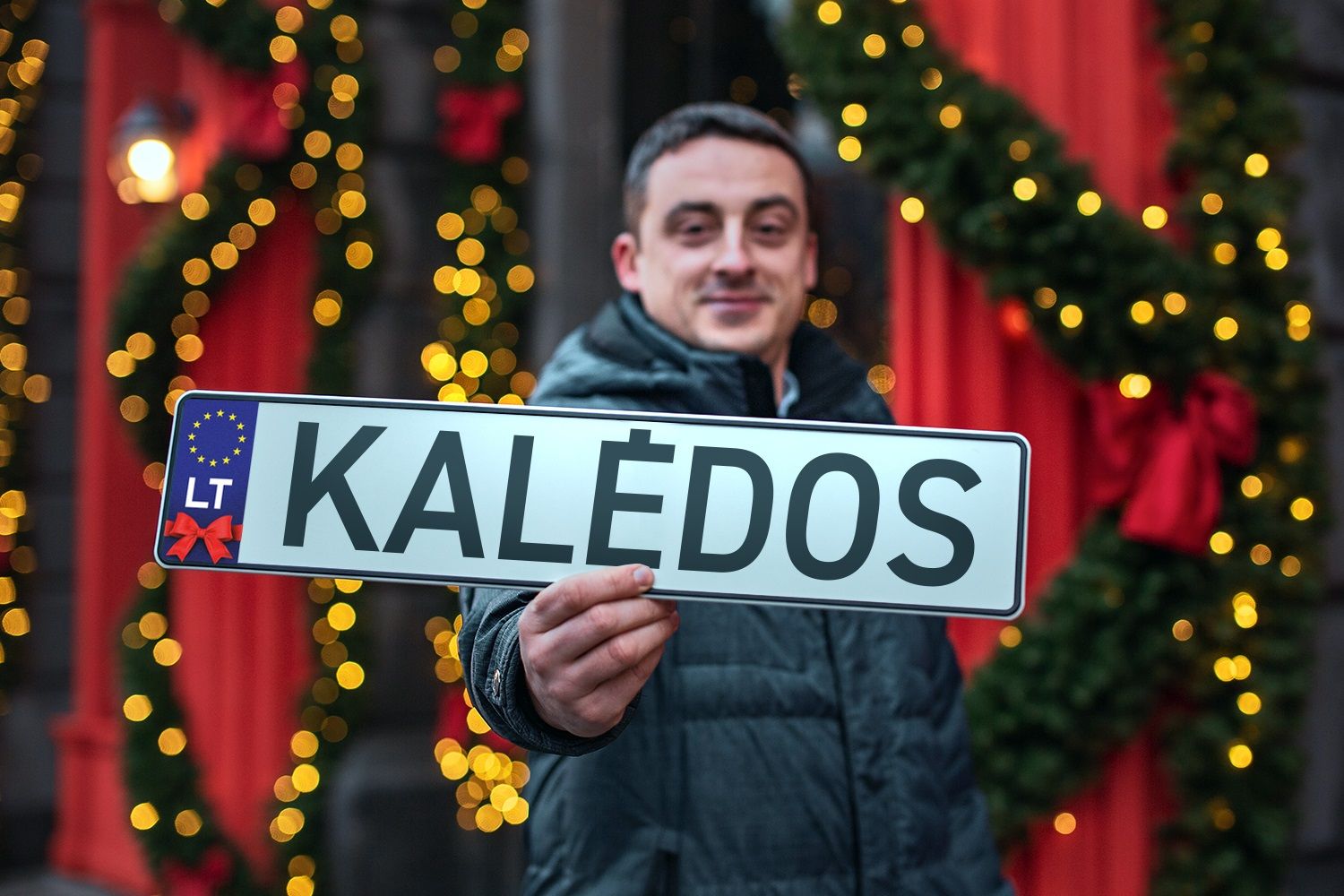 Asmuo laiko lentelę su užrašu „KALĖDOS“, primenančią ES numerio ženklą, fone – kalėdinės dekoracijos.