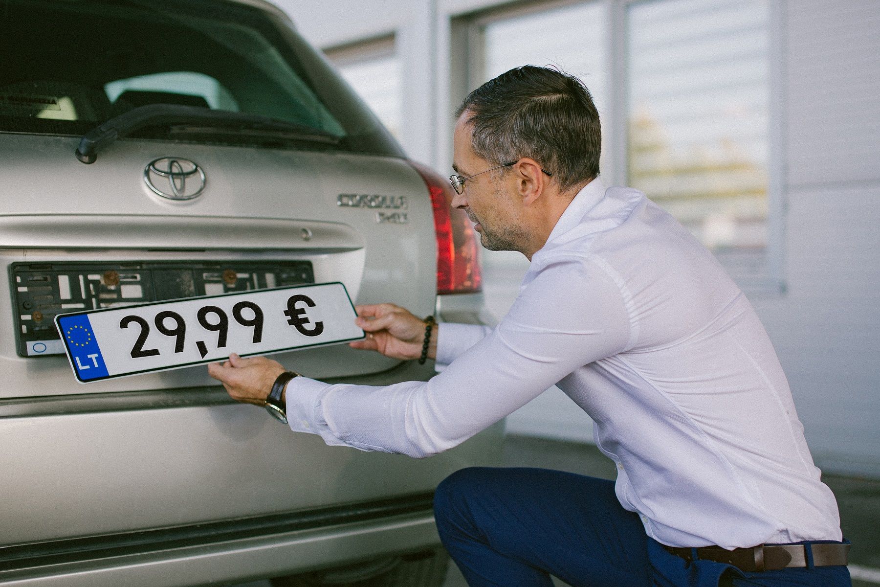 Asmuo su baltais marškiniais ir mėlynomis kelnėmis laiko ženklą „29,99 €“ prie sidabrinės „Toyota“ automobilio galinės dalies su lietuviškais numeriais