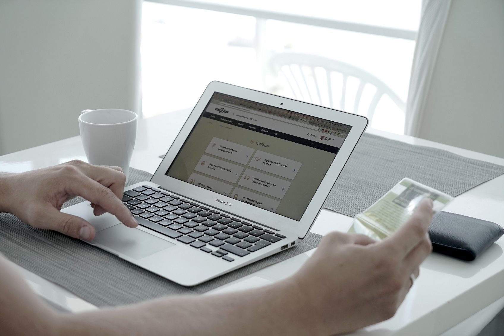 Asmuo prie stalo naudoja „MacBook Air“ kompiuterį, kurio ekrane matomas interneto puslapis su įvairiomis kategorijomis. Kitoje rankoje – išmanusis telefonas. Ant stalo – balta kavos puodelis ir piniginė.