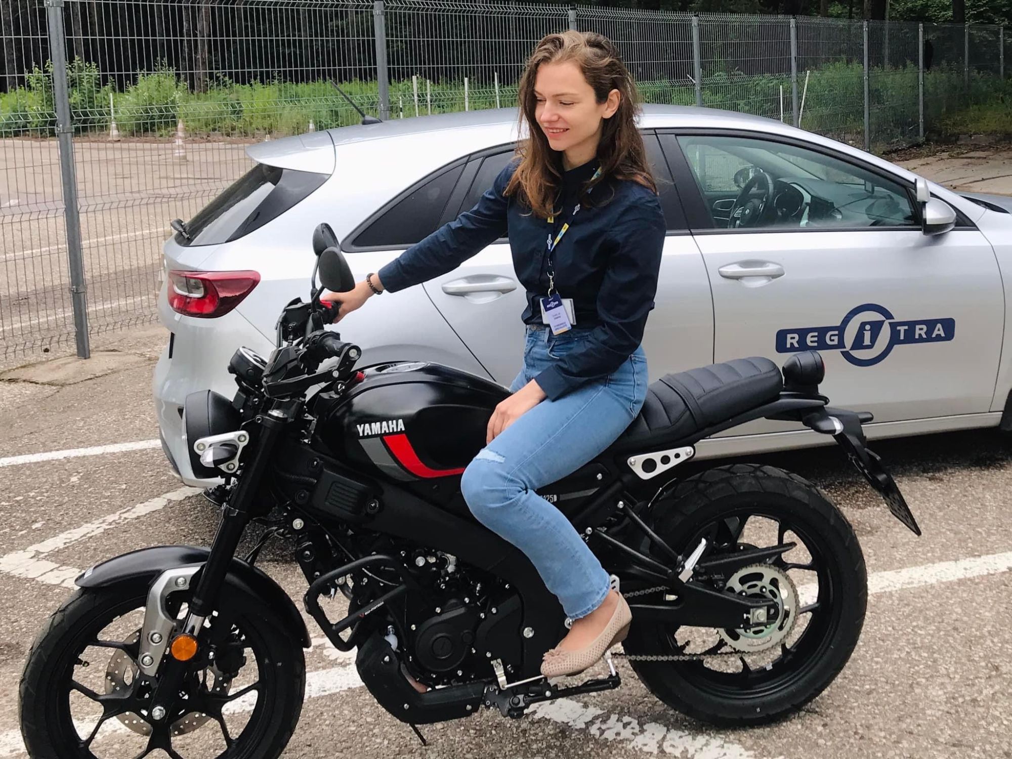 Asmuo sėdi ant juodo „Yamaha“ motociklo lauke, vilki tamsius marškinėlius ir mėlynus džinsus, ant kaklo – pažymėjimas. Fone – sidabrinis automobilis su „REGITRA“ logotipu, stovintis aptvertoje aikštelėje.