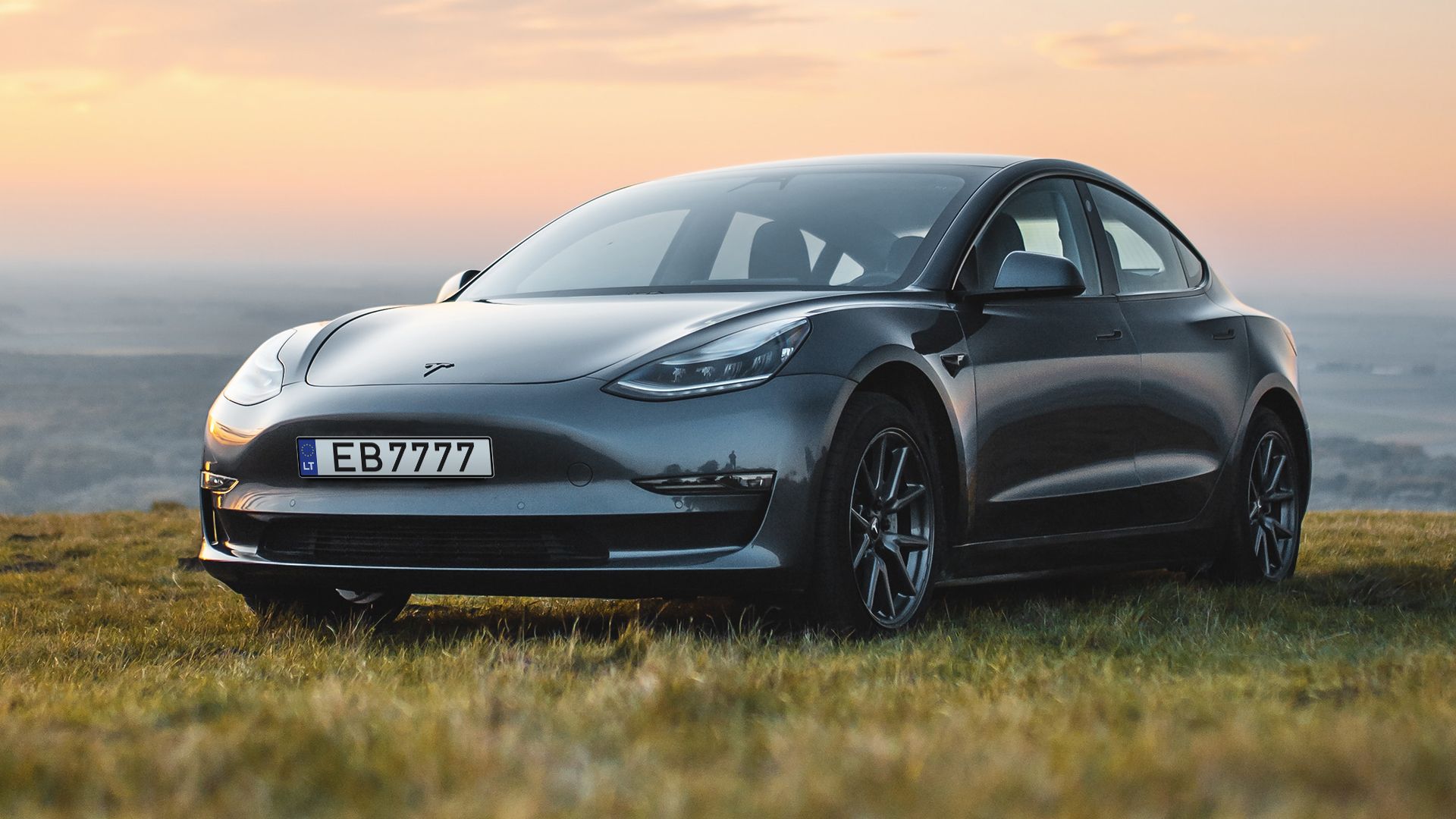 Tamsiai pilkas „Tesla Model 3“ automobilis stovi žolėtame lauke, fone – kalvos ir oranžinis saulėlydžio dangus. Numerio lentelė – „EB7777“.