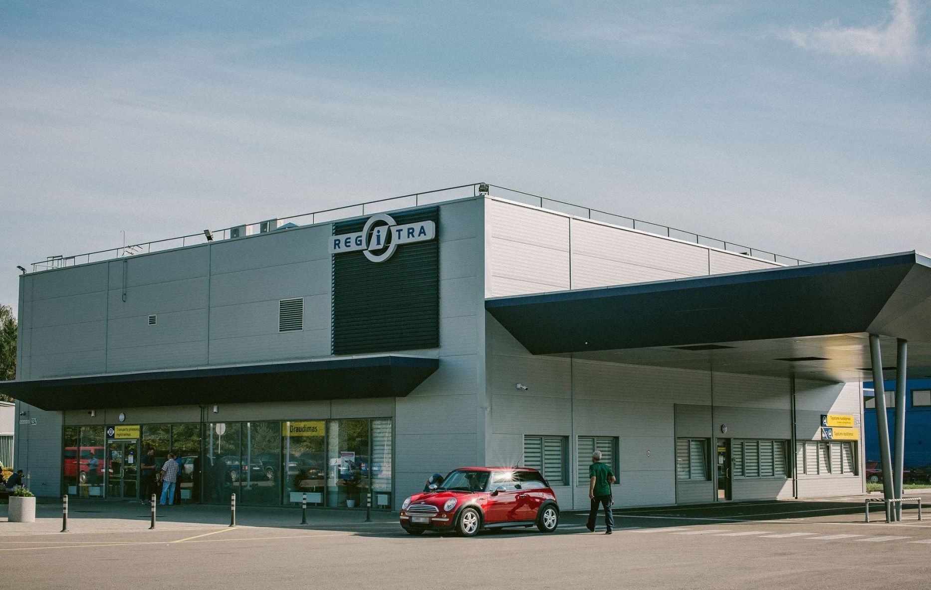 Modernus pastatas su užrašu „REGITRA“. Prie įėjimo matomi žmonės, o priekyje – raudonas „Mini Cooper“ automobilis. Pastatas turi didelius langus ir stogelį virš įėjimo. Giedras dangus.