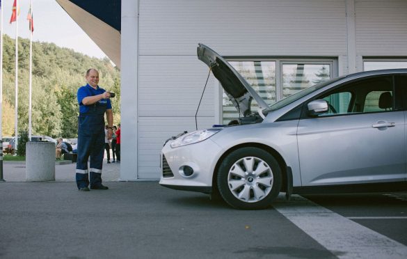Asmuo su mėlyna uniforma stovi šalia atviro kapoto sidabrinio automobilio. Rankoje laiko įrankį ar prietaisą. Fone – pastatas su dideliais langais ir keliomis vėliavomis.