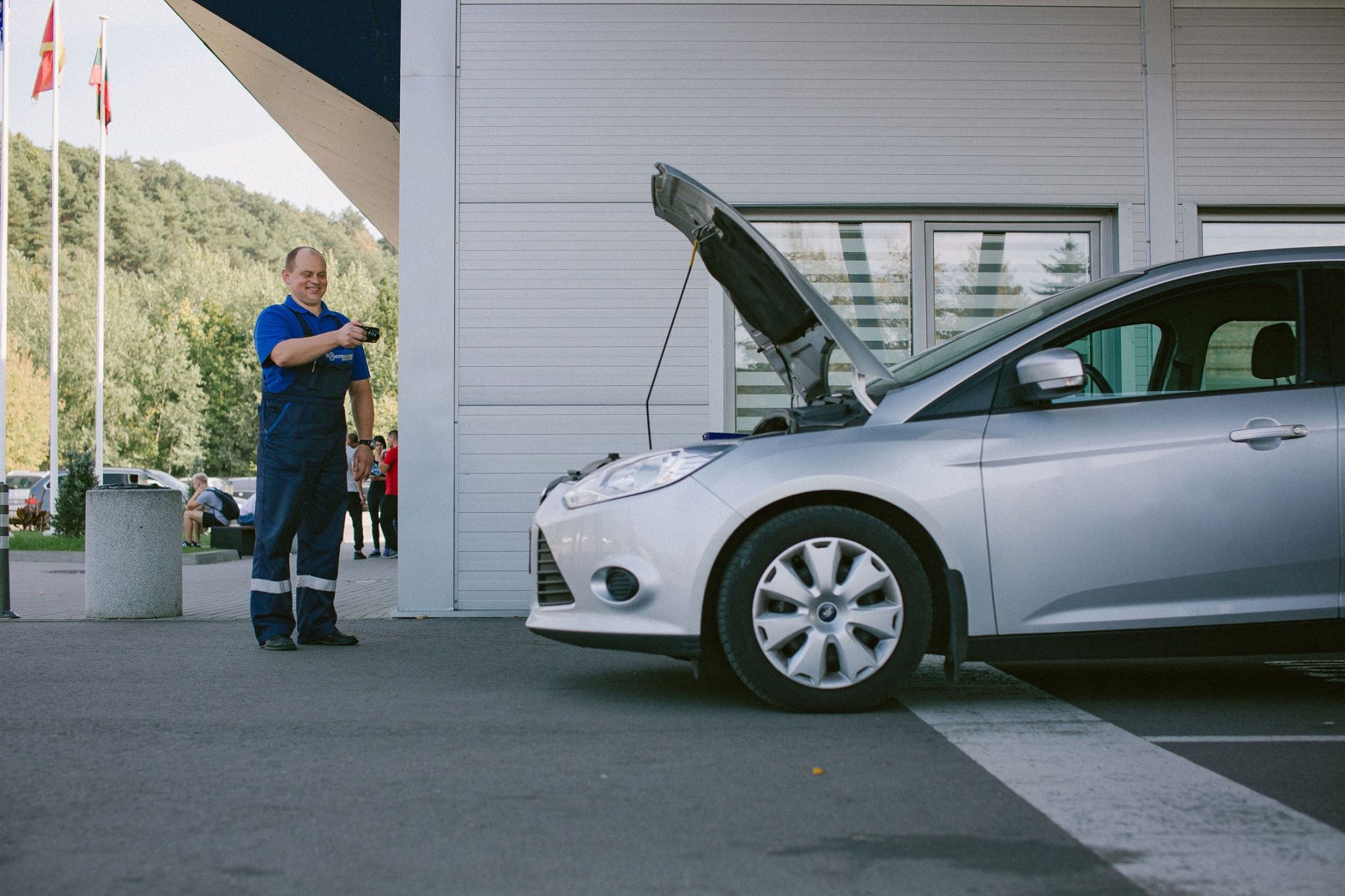 Asmuo su mėlyna uniforma stovi šalia atviro kapoto sidabrinio automobilio. Rankoje laiko įrankį ar prietaisą. Fone – pastatas su dideliais langais ir keliomis vėliavomis.