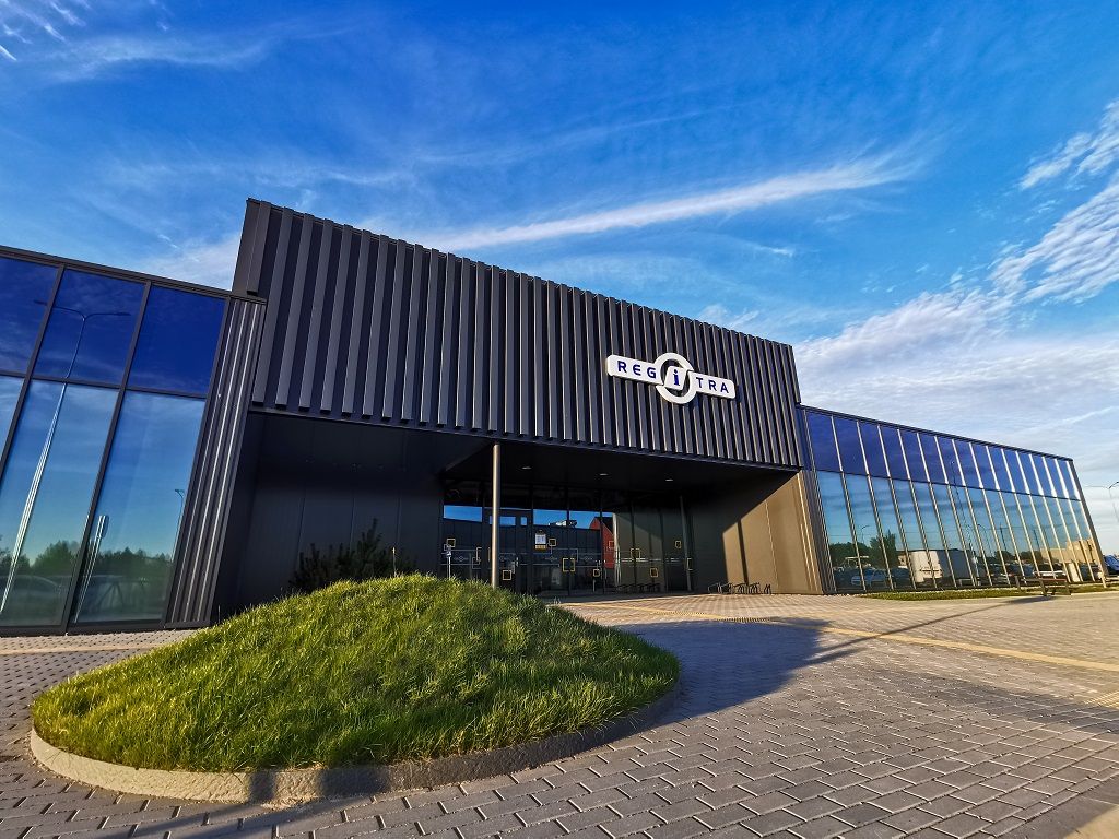 Modernus pastatas su tamsiu, gofruoto metalo fasadu ir dideliais stikliniais langais. Priekyje matomas „REGITRA“ logotipas ir užrašas. Prie pastato – nedidelė žolės kalvelė, viršuje – giedras dangus su plonais debesimis.
