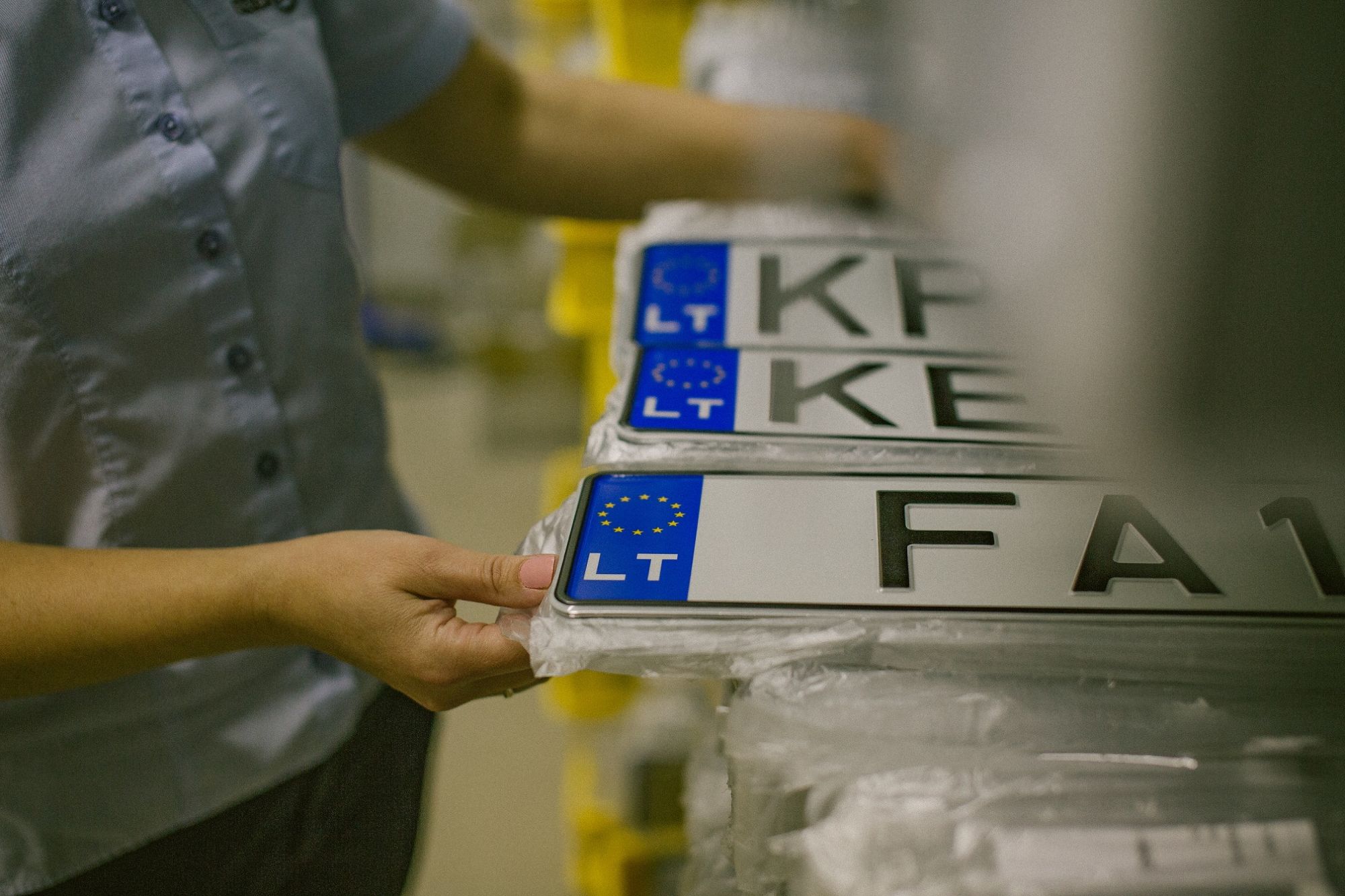Asmuo laiko krūvą lietuviškų transporto priemonių numerio lentelių su ES vėliava ir žyma „LT“. Matomi numeriai: „KP“, „KE“ ir „FA1“.
