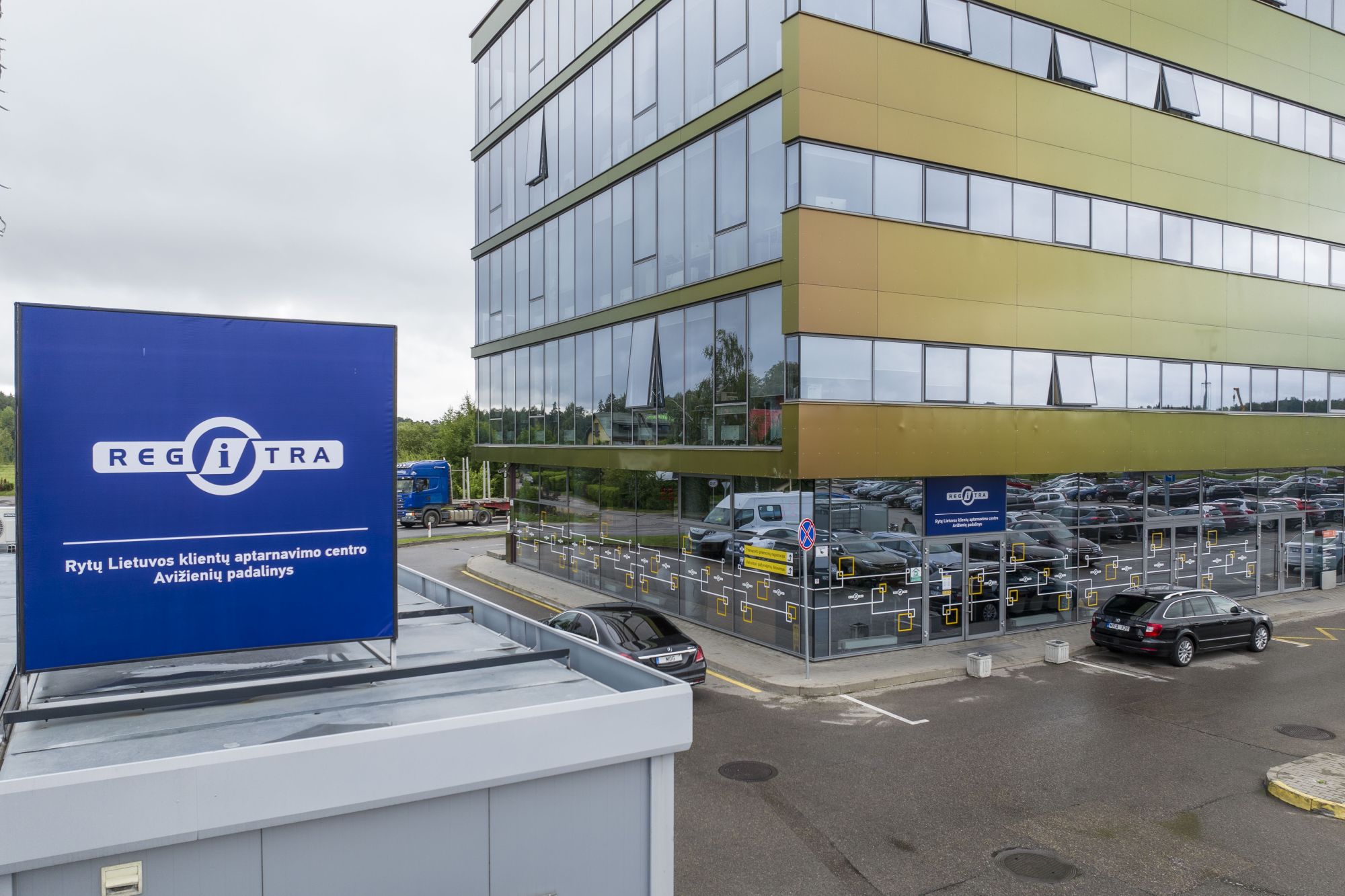 Modernus biurų pastatas su dideliais stikliniais langais ir automobilių stovėjimo aikštele priekyje. Matomas mėlynas ženklas su užrašu „REGITRA“ ir tekstu „Rytų Lietuvos klientų aptarnavimo centro Avižienių padalinys“