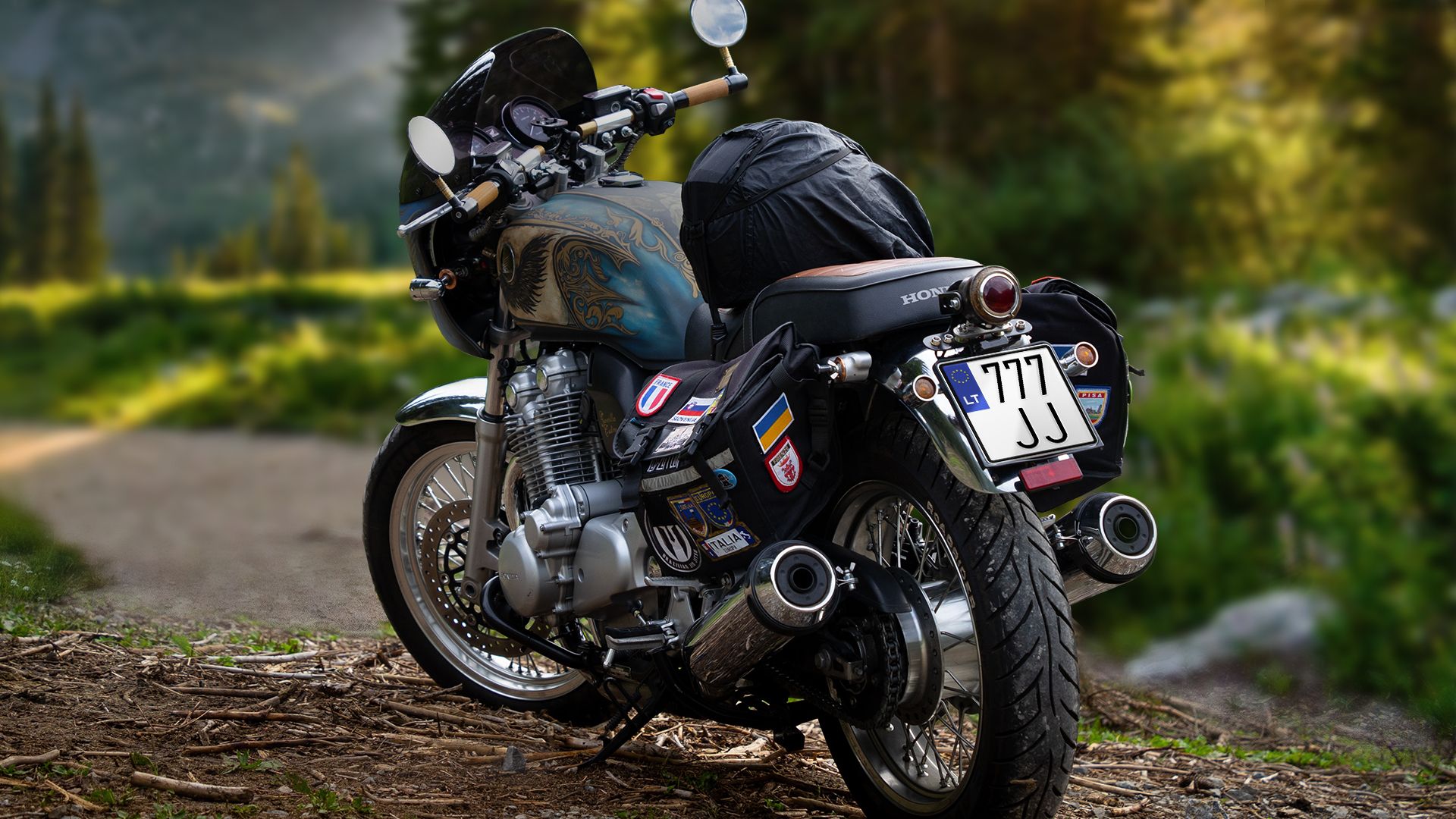 Motociklas stovi miško keliuke. Ant šoninių krepšių – įvairūs lipdukai su vėliavomis ir logotipais, gale pritvirtintas didelis juodas krepšys. Numerio lentelė – „777 JJ“. Fone – medžiai ir saulės apšviesta žaluma.