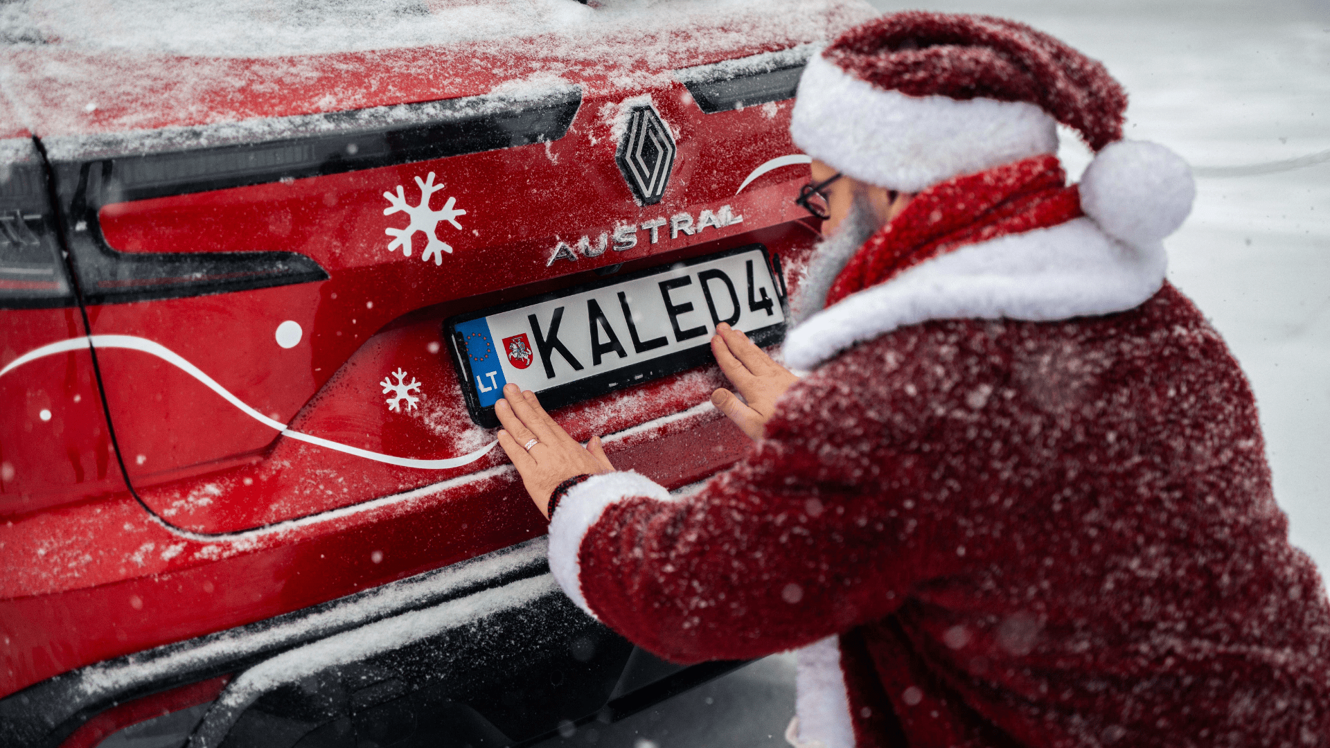 Asmuo su Kalėdų Senelio kostiumu tvirtina numerio lentelę „KALED4“ prie raudono „Renault Austral“ automobilio. Automobilis papuoštas snaigių lipdukais, aplink – sniegas.
