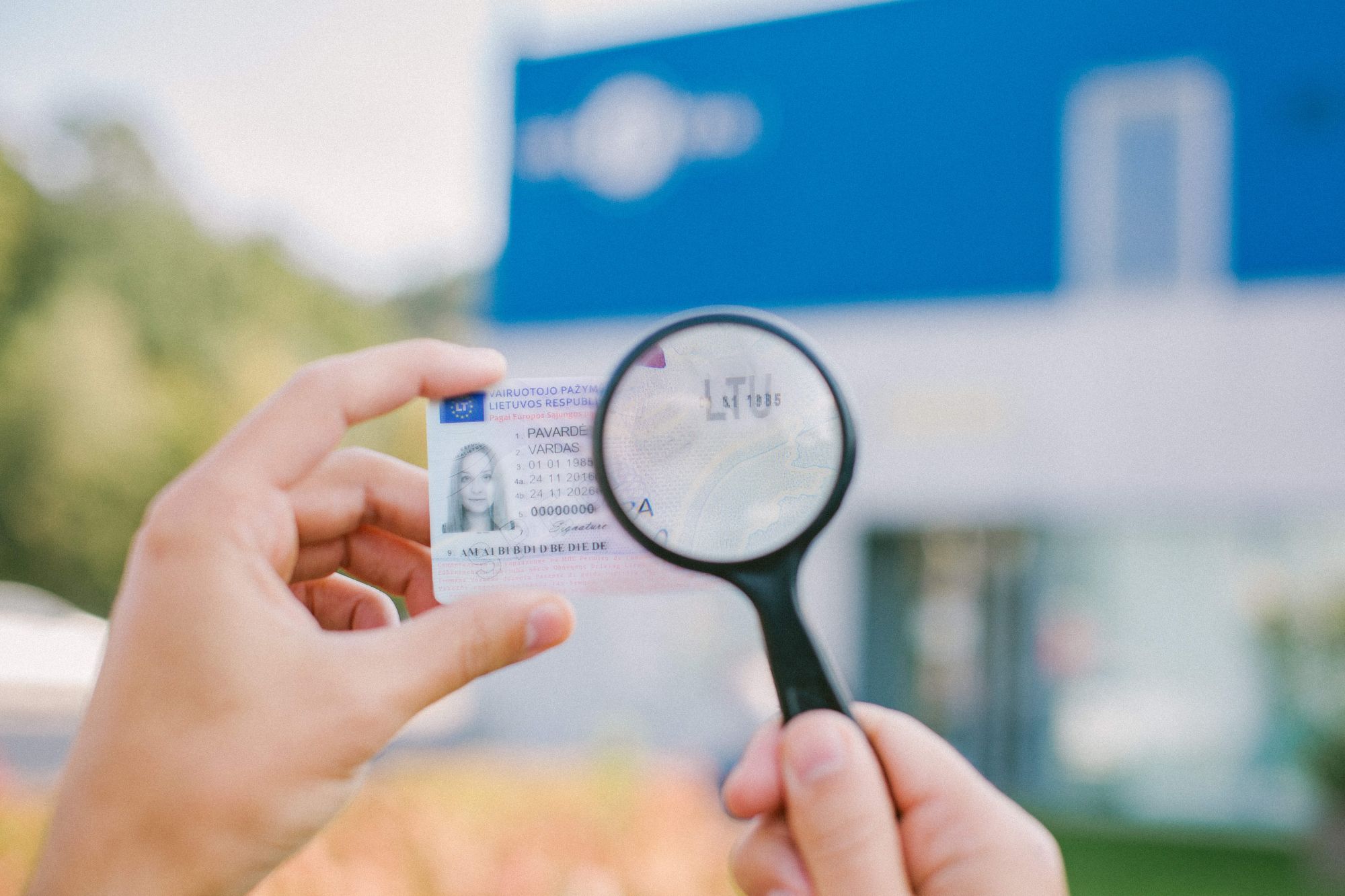 Asmuo laiko padidinamąjį stiklą, kad įžiūrėtų detales ant asmens tapatybės kortelės. Fone matomas neryškus mėlynos ir baltos spalvų pastatas lauke.