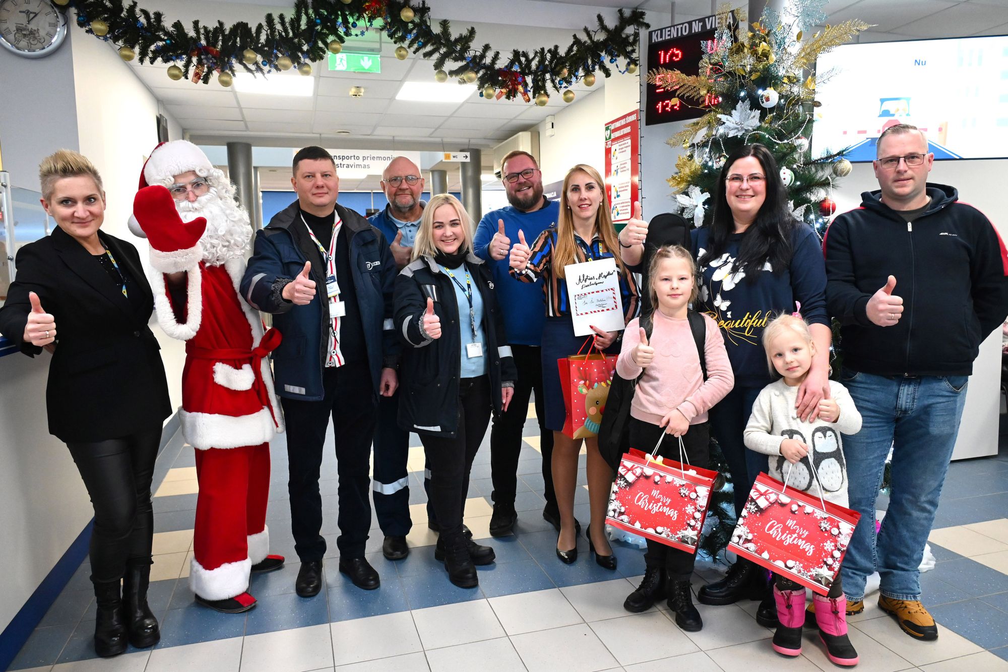 Grupė žmonių stovi koridoriuje, papuoštame kalėdinėmis girliandomis ir eglute. Vienas asmuo apsirengęs Kalėdų Seneliu, keli rodo nykščius į viršų, kai kurie laiko dovanų maišelius, vienas – pažymėjimą ar plakatą. Fone matomas elektroninis ekranas su tekstu ir skaičiais.