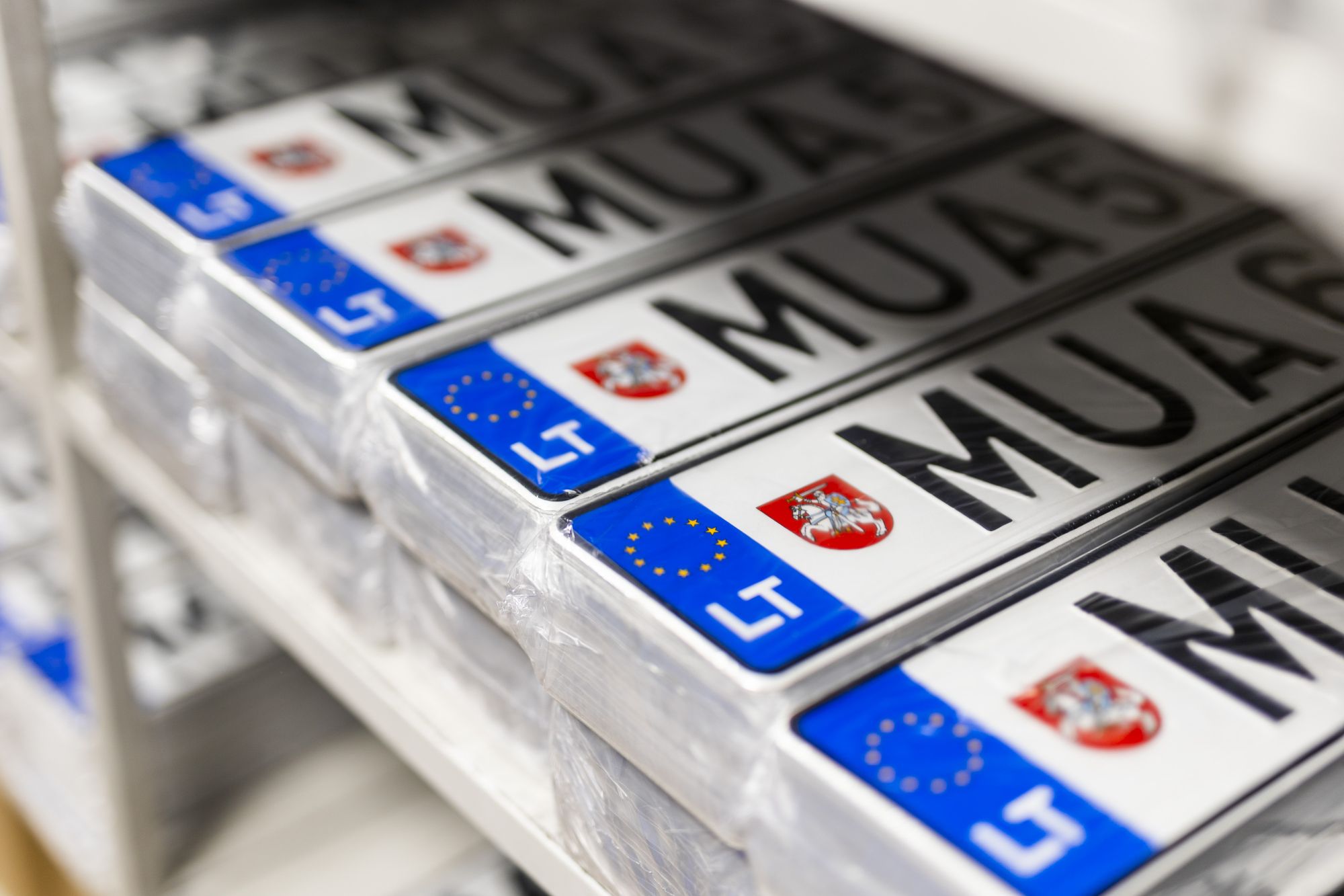 Lietuviškų transporto priemonių numerio lentelių krūva lentynoje. Ant lentelių – raidės „MUA“, skaičiai, Lietuvos herbas ir ES vėliava su žyma „LT“.