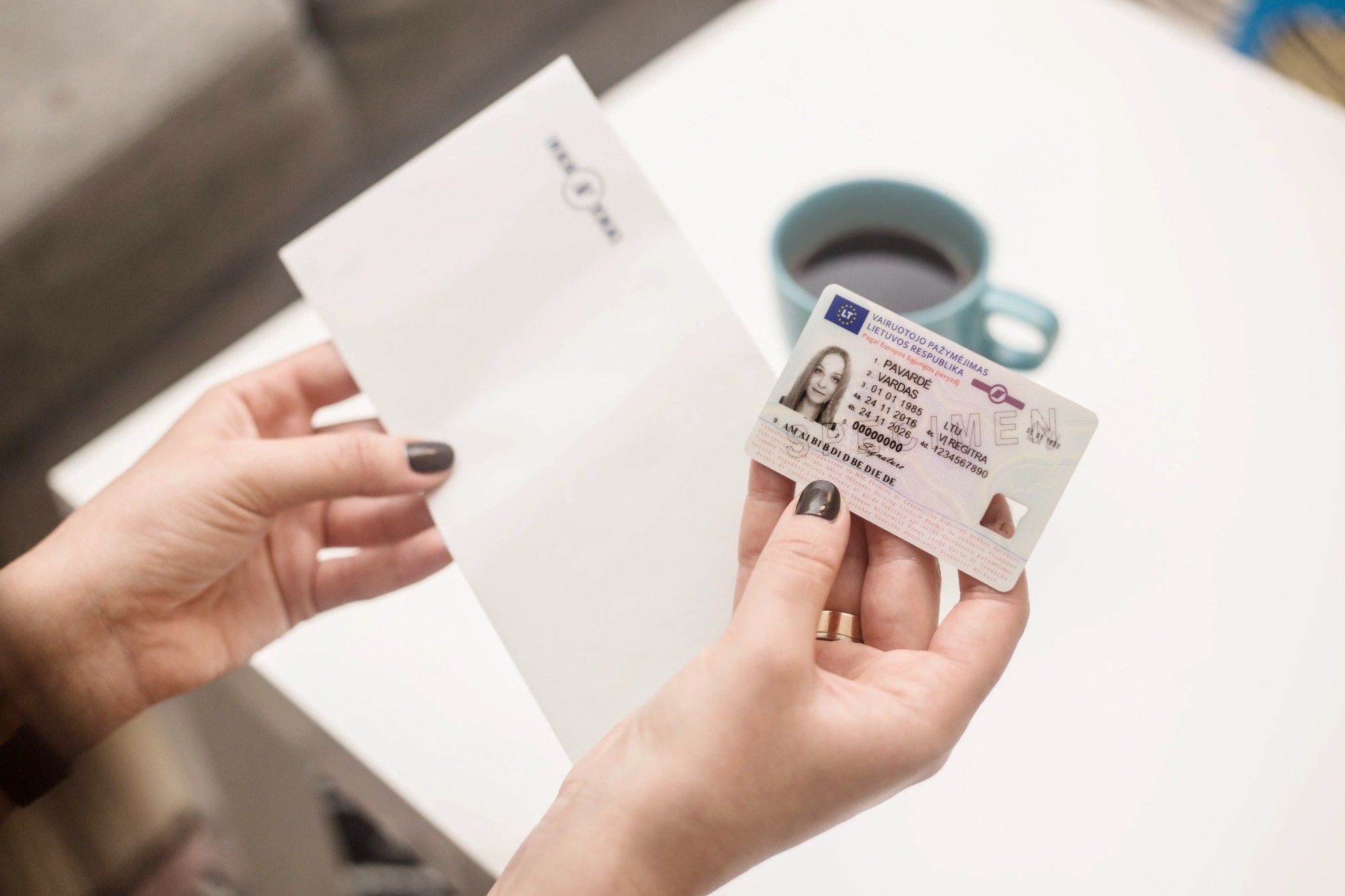 Asmuo laiko asmens tapatybės kortelę vienoje rankoje ir baltą voką – kitoje. Fone ant stalo matomas mėlynas puodelis su kava.