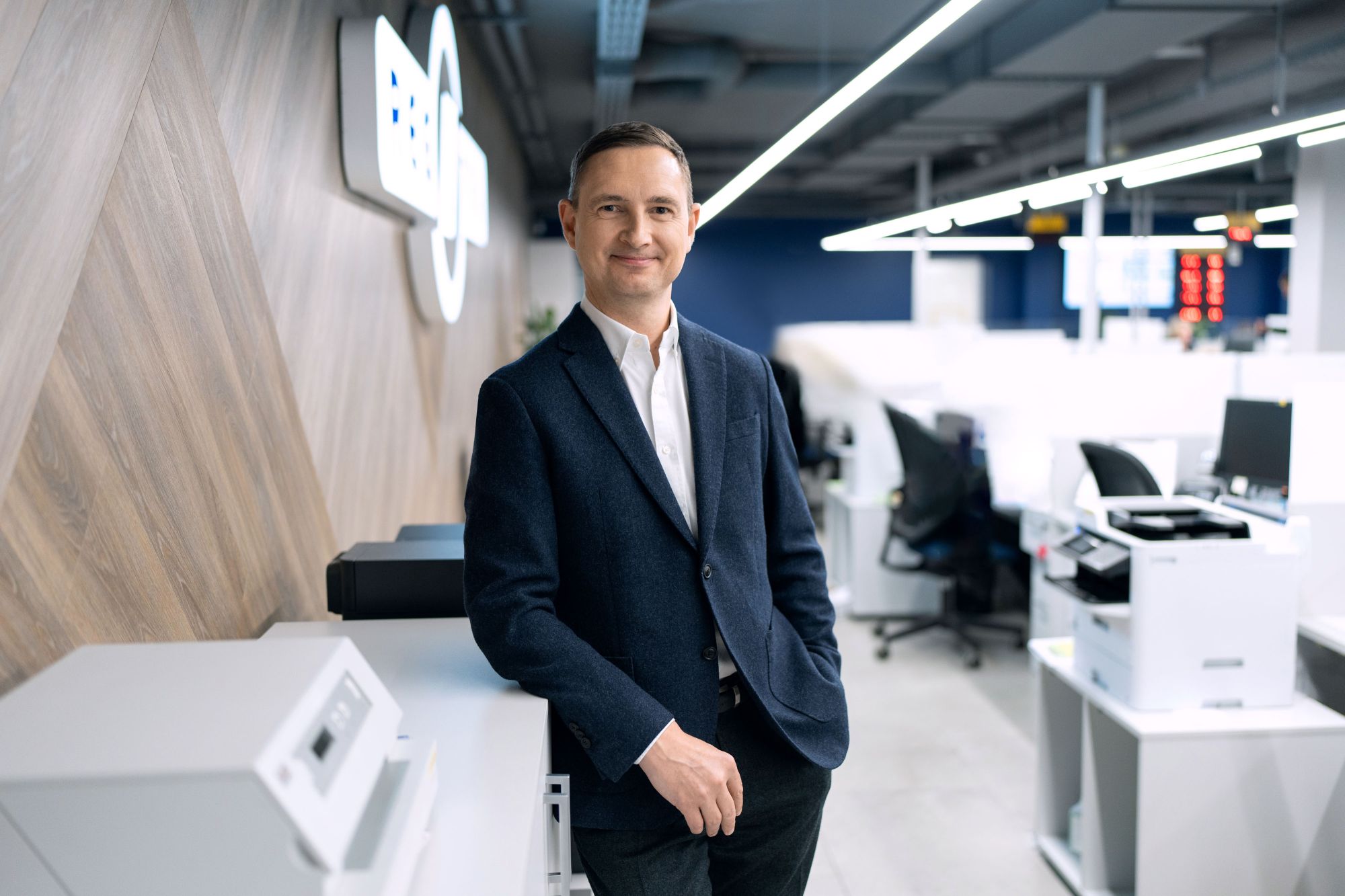 Šioje nuotraukoje matomas asmuo, vilkintis tamsų švarką, baltus marškinius ir turintis nosinaitę švarko kišenėje. Fone – stiklinė siena su geometriniais raštais, kairėje pusėje matomas „REGITRA“ logotipas.