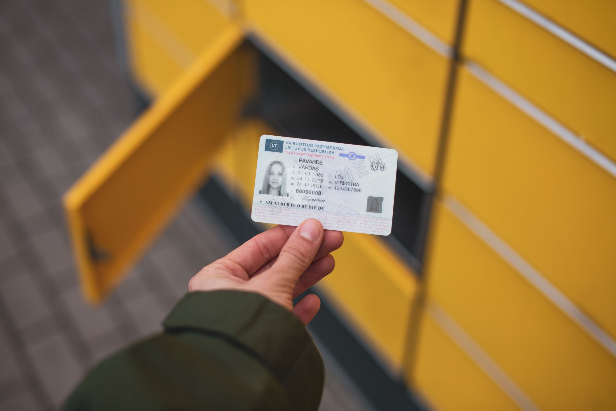 Nuotraukoje matoma ranka, laikanti identifikacinę kortelę (galimai vairuotojo pažymėjimą arba asmens tapatybės kortelę) priešais geltonų skyrelių sistemą, panašią į paštomatą ar siuntų terminalą. Vienas iš skyrelių yra atidarytas.