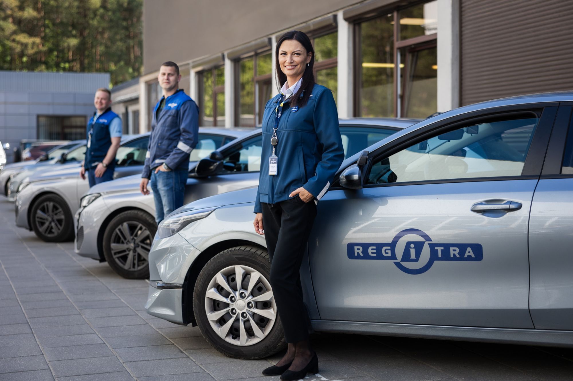 Nuotraukoje matomi trys asmenys mėlynais uniforminiais drabužiais, stovintys šalia automobilių eilės, ant kurių šonų matomas „REGITRA“ logotipas.