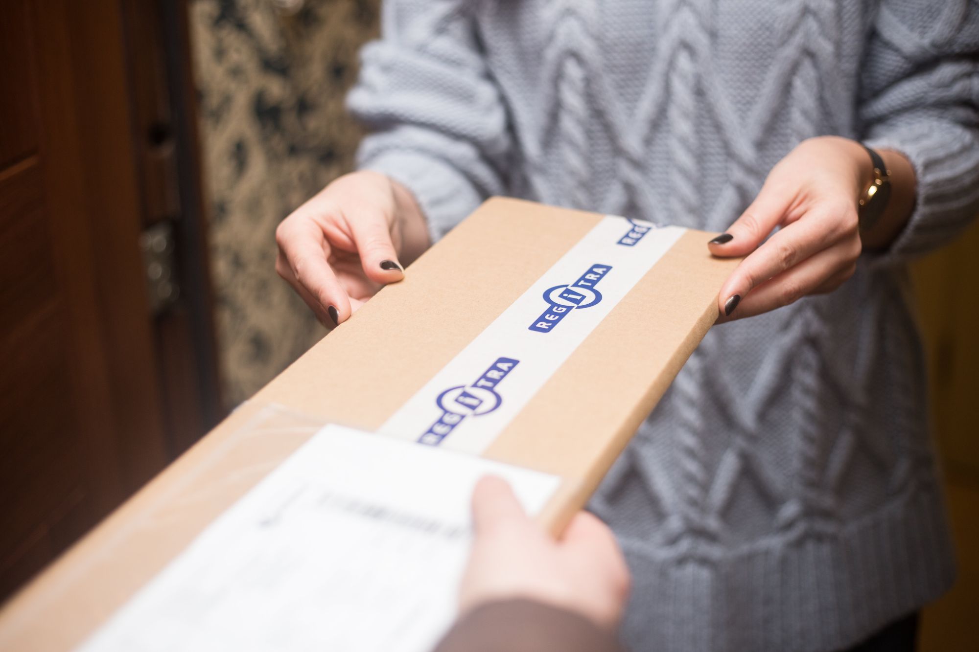 Asmuo, vilkintis pilką megztinį, kuris gauna kartoninę siuntą iš kito asmens. Ant siuntos yra balta etiketė su mėlynu tekstu ir logotipais, kas rodo, kad tai gali būti oficialus ar registruotas siuntinys.