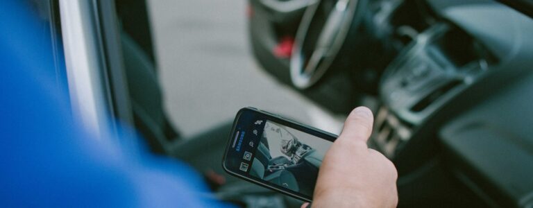 Nuotraukoje matomas asmuo, laikantis išmanųjį telefoną ir fotografuojantis automobilio saloną. Fokusas yra nukreiptas į telefono ekraną, kuriame matomas fotografuojamas vaizdas. Fone matomi vairas ir prietaisų skydelis.