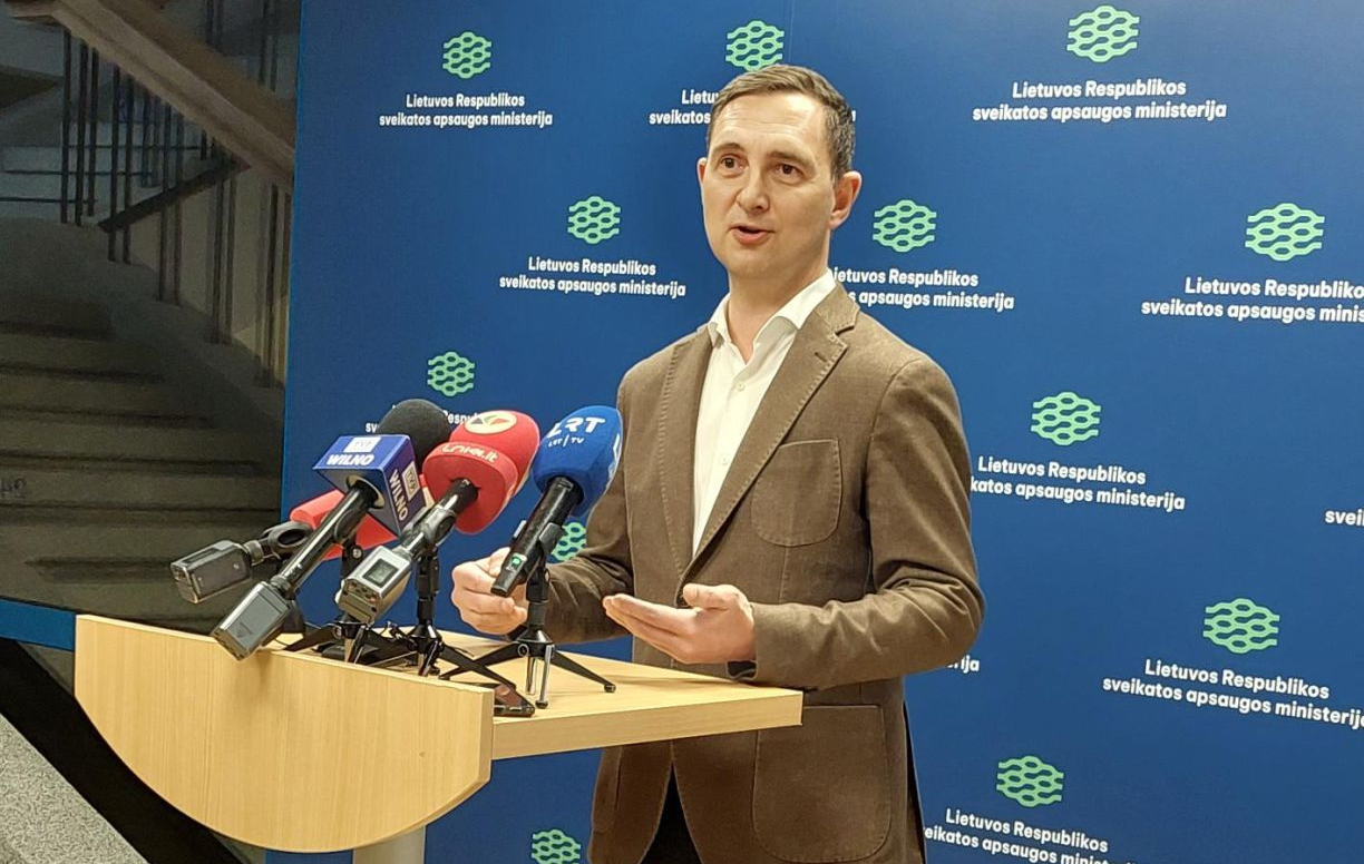 Asmuo, stovintis prie pakylos su keliais mikrofonais, priklausančiais skirtingoms naujienų agentūroms. Fone – mėlynas fonas su pakartotu tekstu.