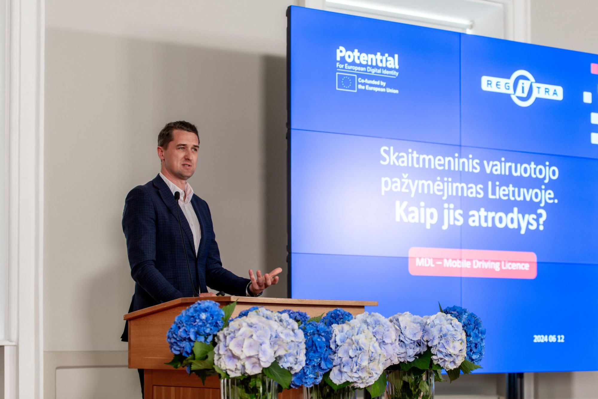 Nuotraukoje matomas asmuo, stovintis prie pakylos, priešais kurią yra mėlynų ir baltų gėlių kompozicija. Už asmens yra didelis ekranas, kuriame rodomas pristatymo skaidrė su tekstu lietuvių kalba.